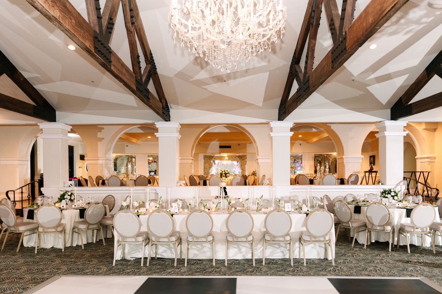 An elegant white wedding reception ballroom with crystal chandeliers and exposed wooden beams.
