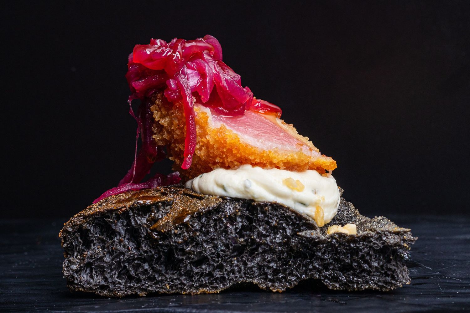 Gourmet black bread sandwich with pink protein and creamy spread on dark background.