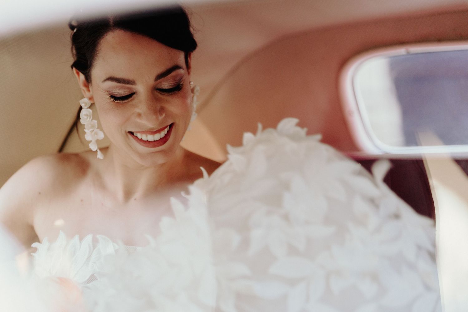 Candid shot of bride in white dress sitting in classic car showing joyful expression.