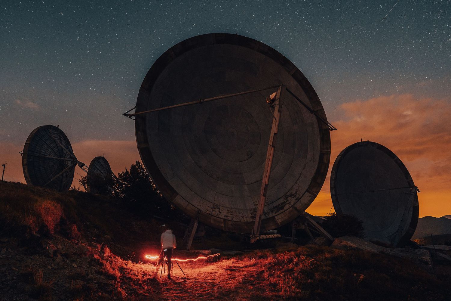 Large satellite dishes silhouetted against sunset sky with figure illuminated by red light.