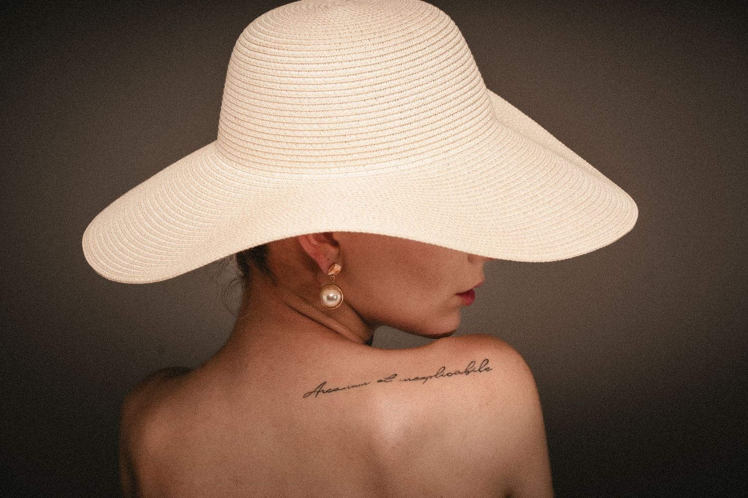 Minimalist portrait of person wearing white sun hat with script tattoo visible on shoulder.
