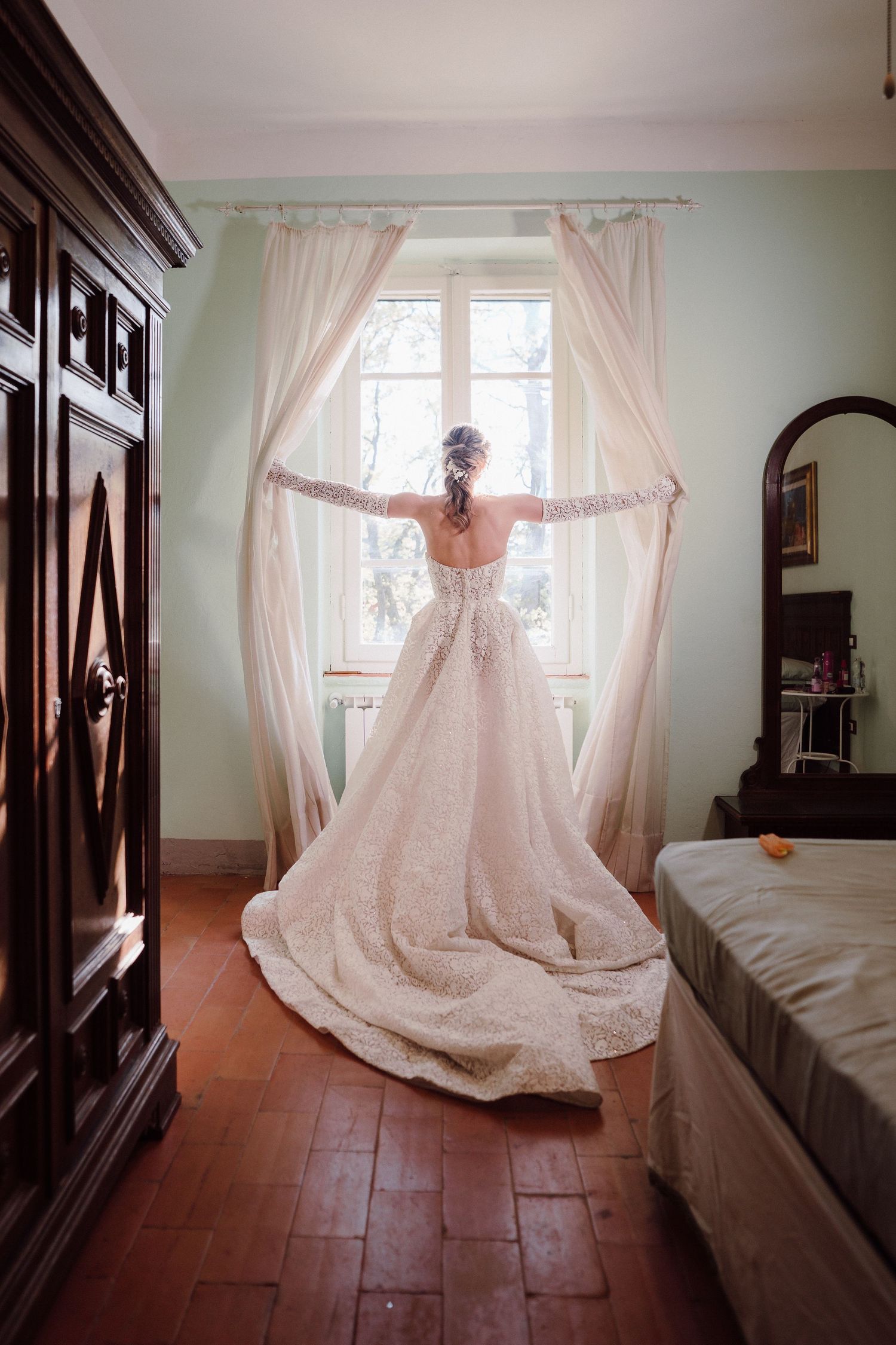 Back view of bride in flowing lace gown standing at window with sheer curtains in sunlit room.