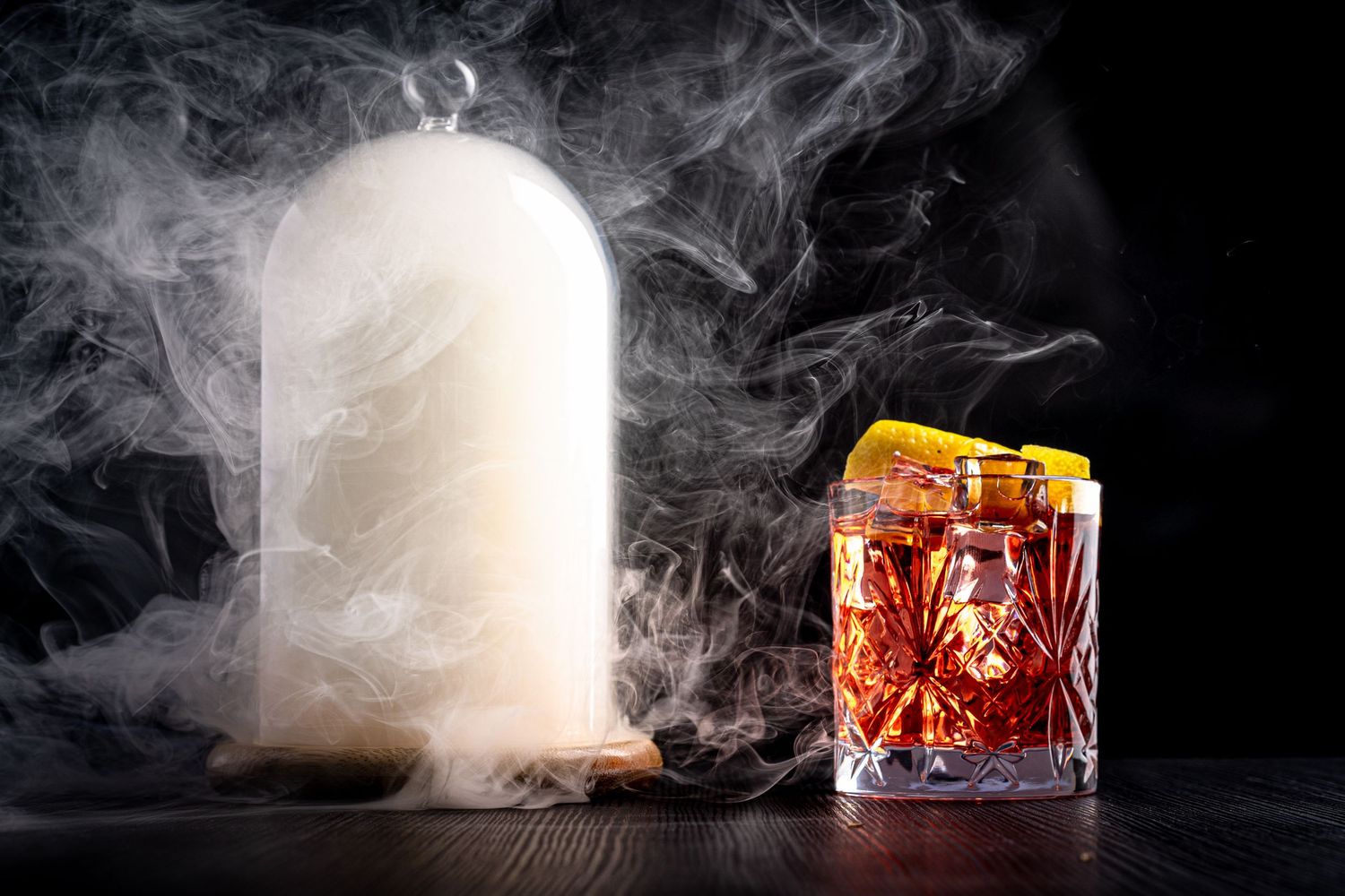 Smoky cocktail presentation with glass cloche and crystal tumbler of amber spirits.