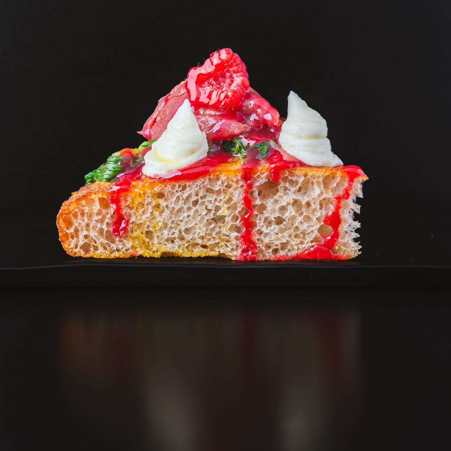 Strawberry cheesecake slice with whipped cream and red sauce drizzle on dark surface.