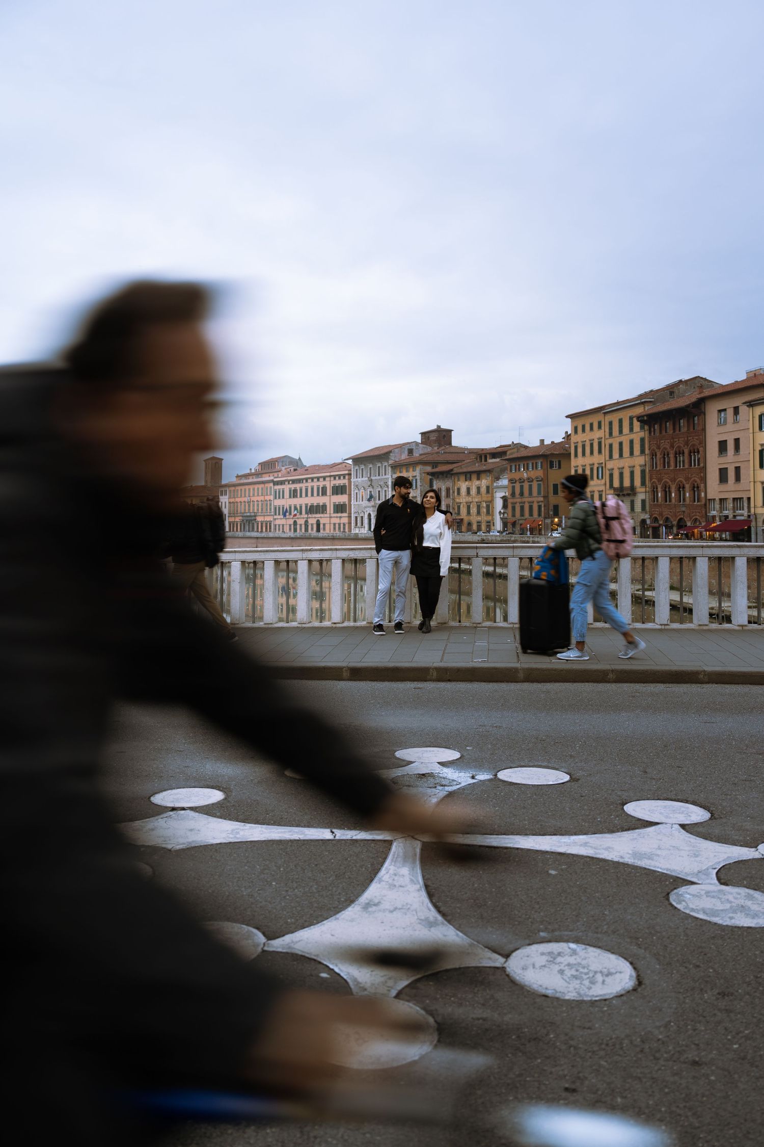A blurred figure passes by as people stand on a bridge in an Italian city.