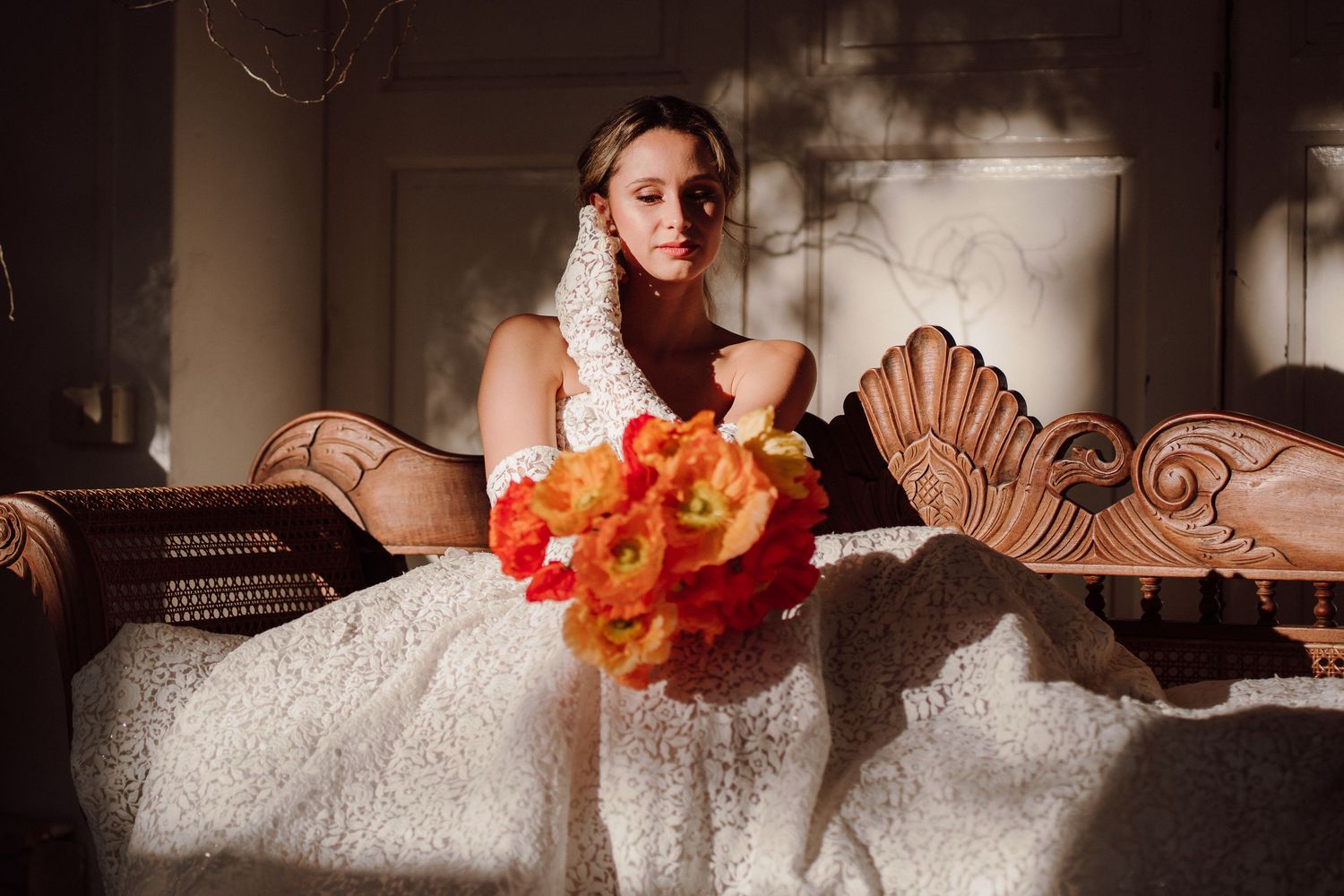Bride in lace dress holding vibrant orange bouquet in dramatic indoor setting.