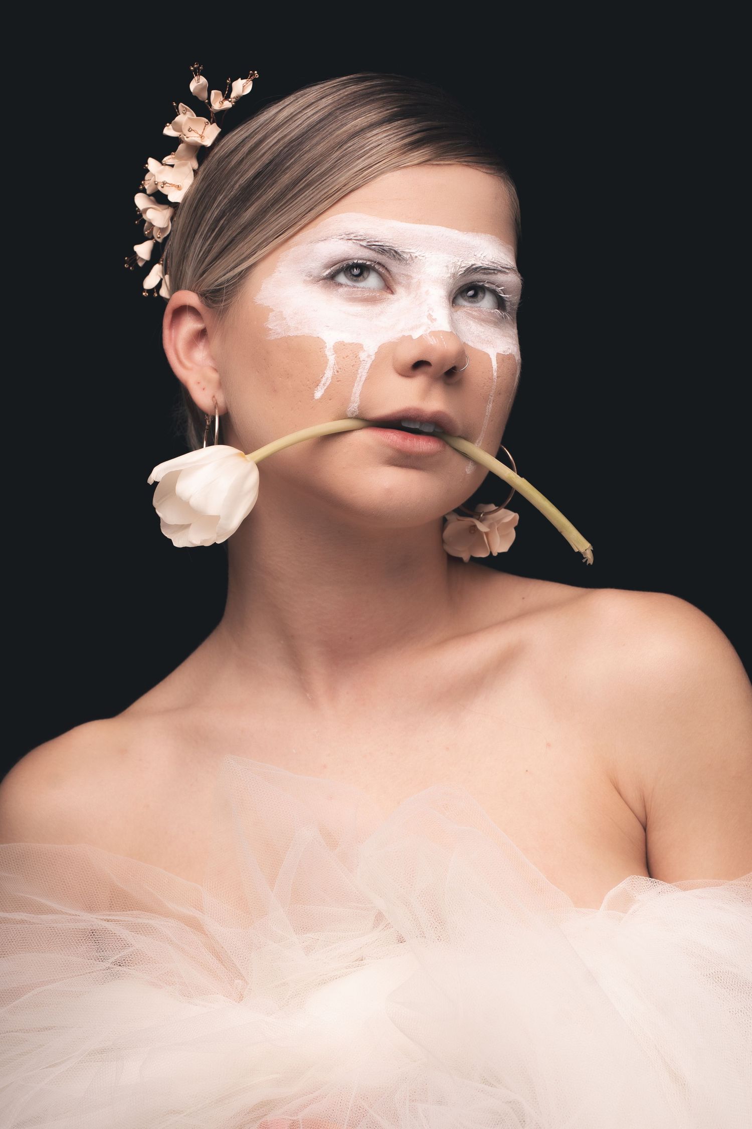 An artistic portrait featuring a model adorned with delicate flowers and white face paint against dark background.
