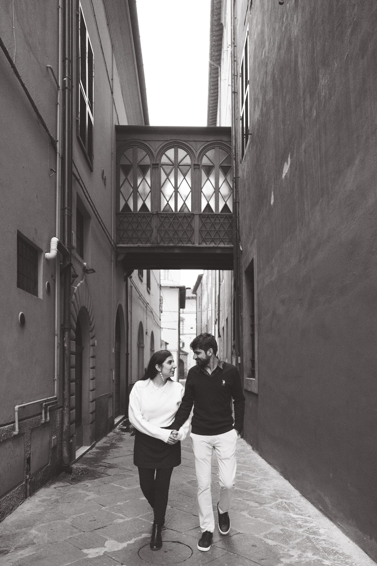 A couple walks together through a narrow alley with an ornate bridge overhead.
