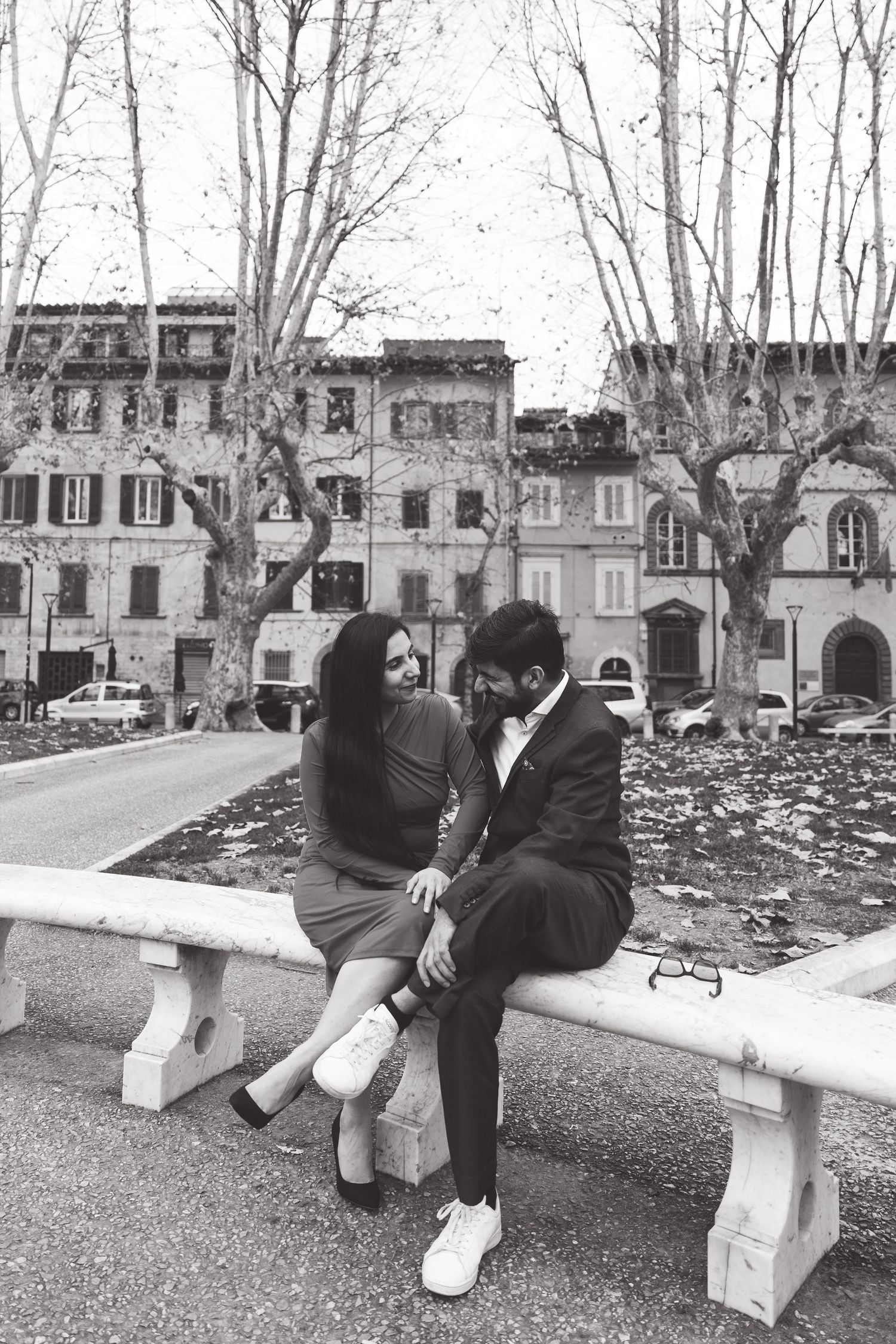 A romantic black and white photo of a couple sitting on a stone bench in an Italian plaza.