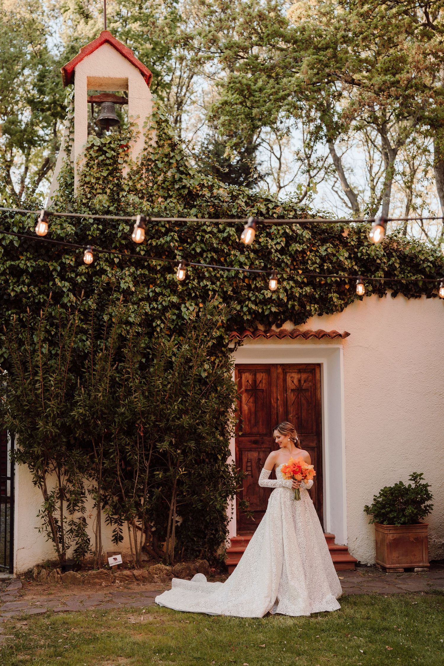 Sunset wedding portrait with string lights and bride in white gown against Spanish-style chapel exterior.