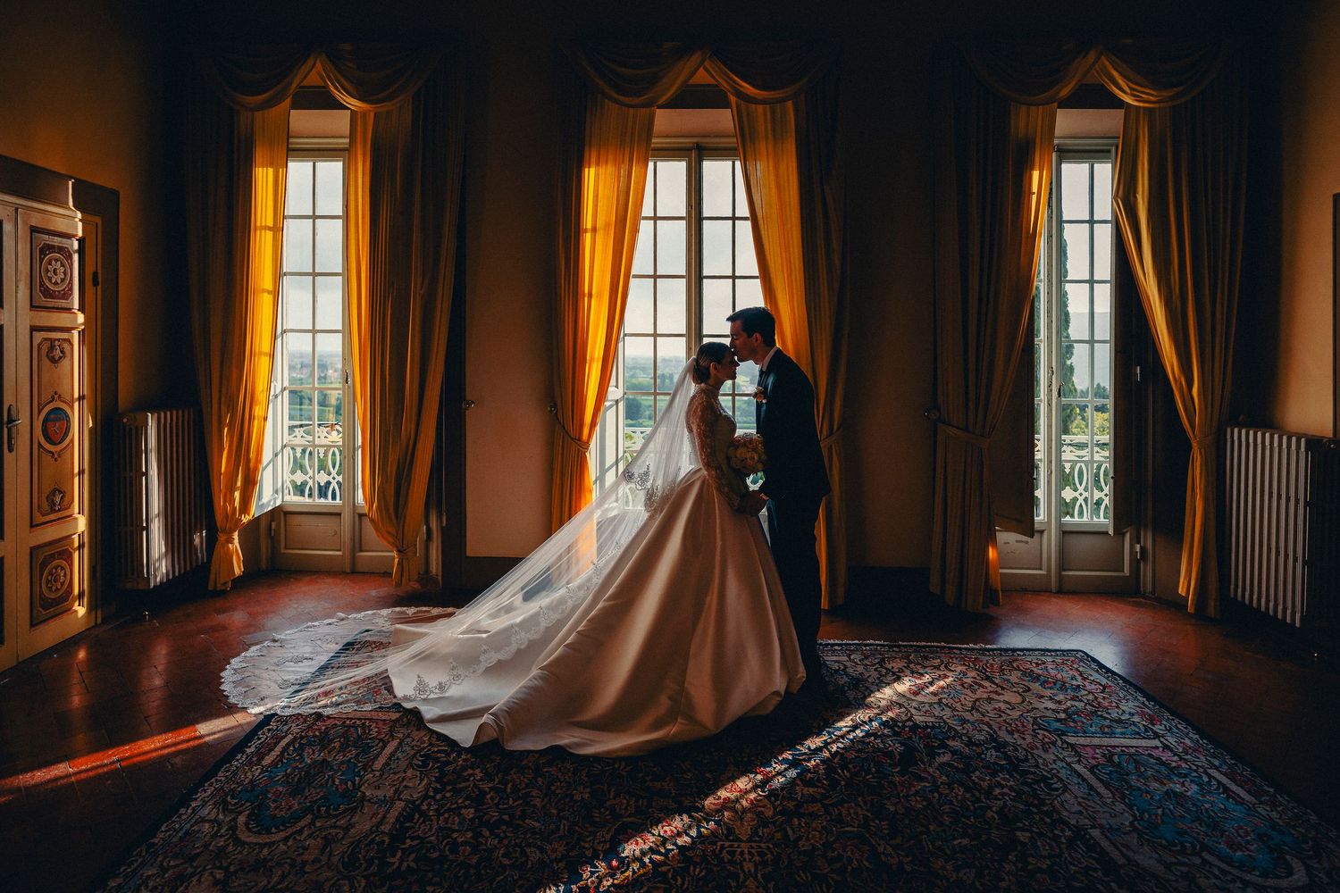 A couple in wedding attire embraces in front of three tall windows in an elegant vintage room with warm lighting.