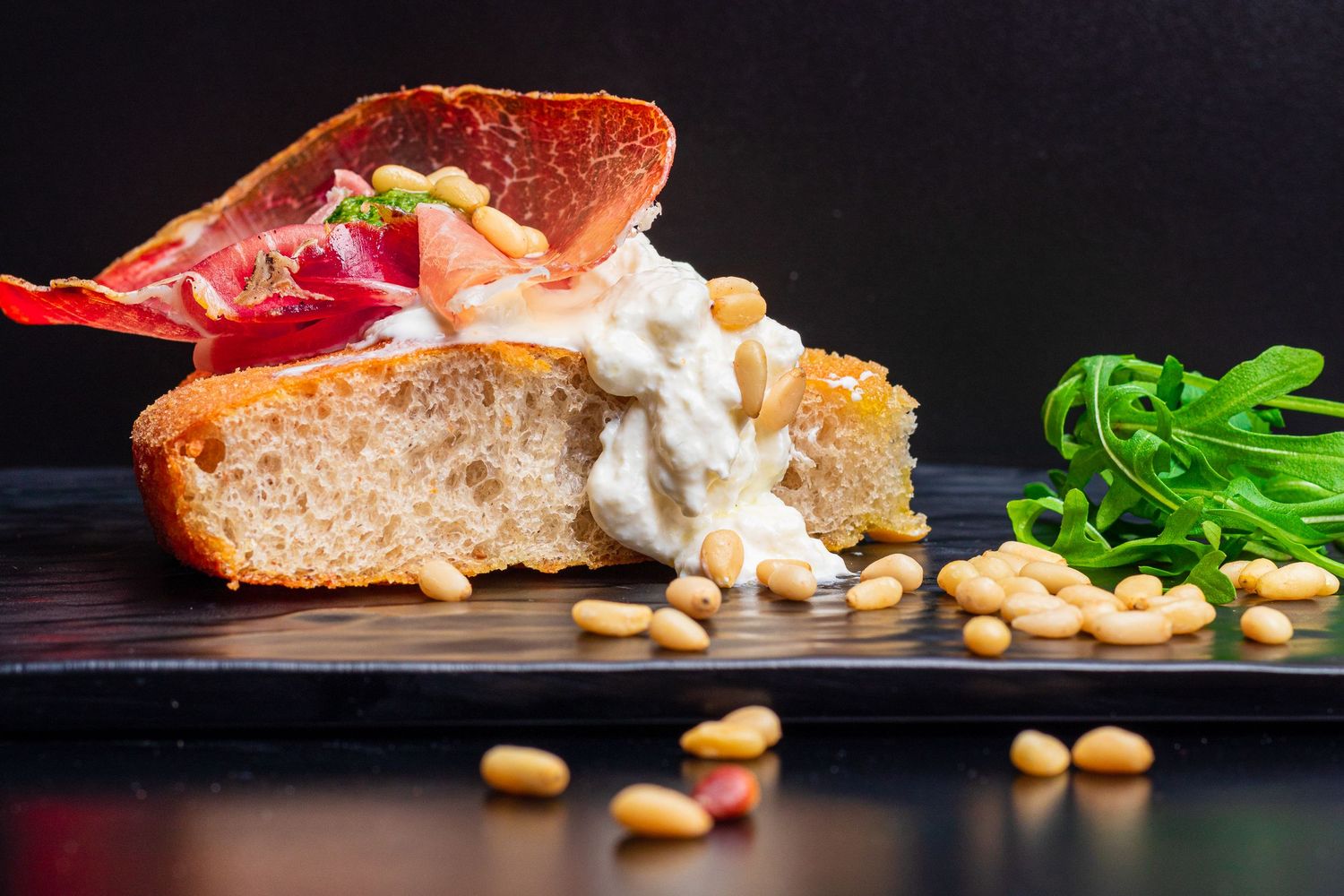 Rustic sandwich with prosciutto, creamy spread and scattered pine nuts on dark surface.