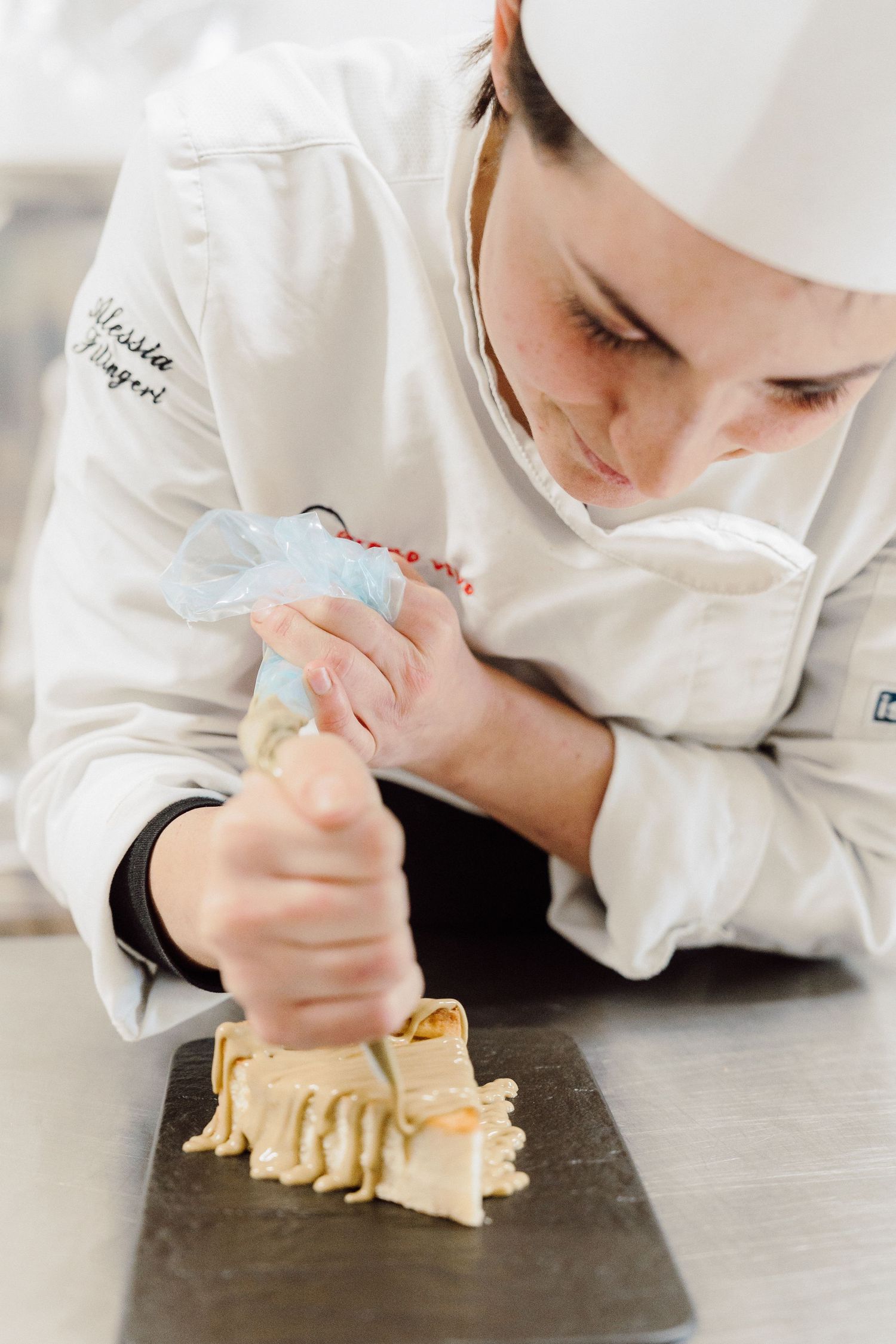 Chef in white uniform piping decorative cream onto a pastry in professional kitchen.