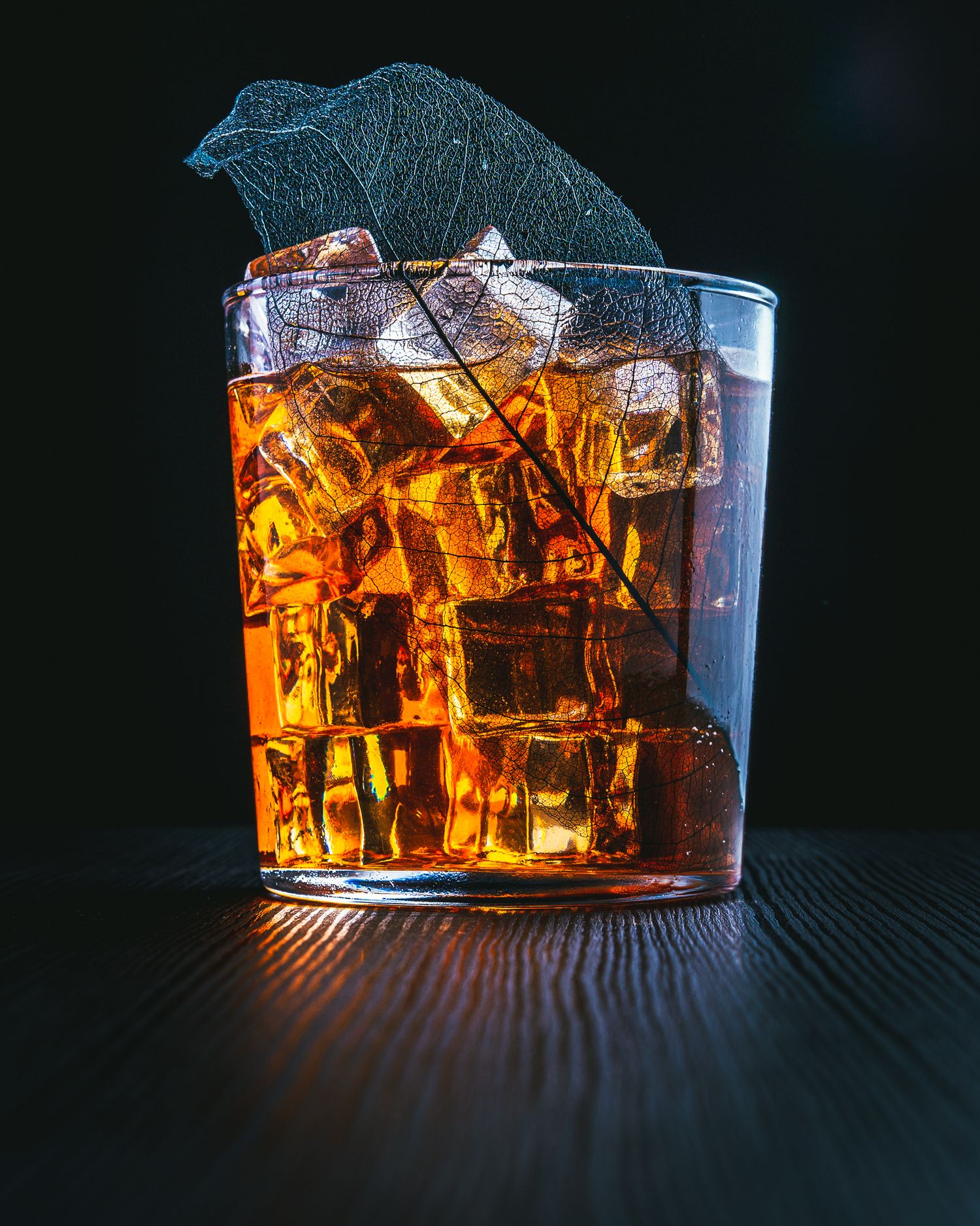 Glass of iced bourbon or whiskey with dramatic lighting on wooden surface.
