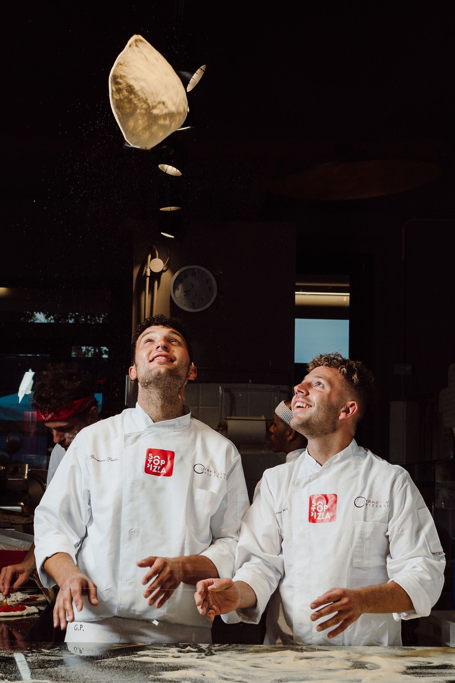 Chefs in white uniforms tossing pizza dough in a restaurant kitchen.