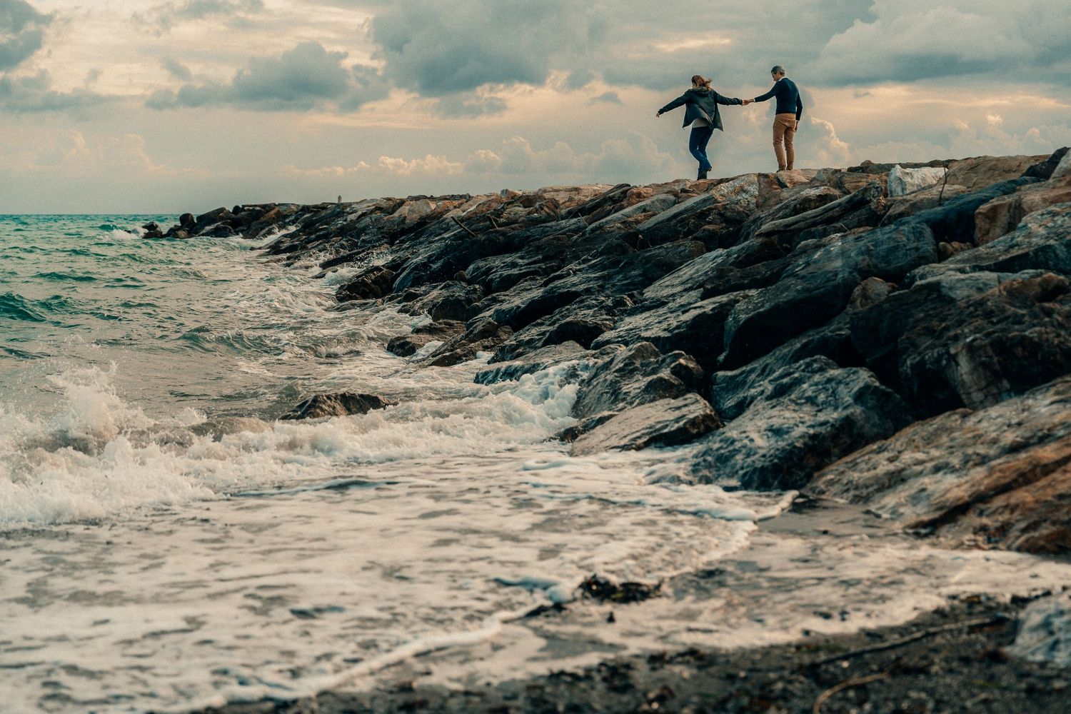 Two silhouettes leap across rocky coastal outcrop with dramatic waves and cloudy sky in the background.