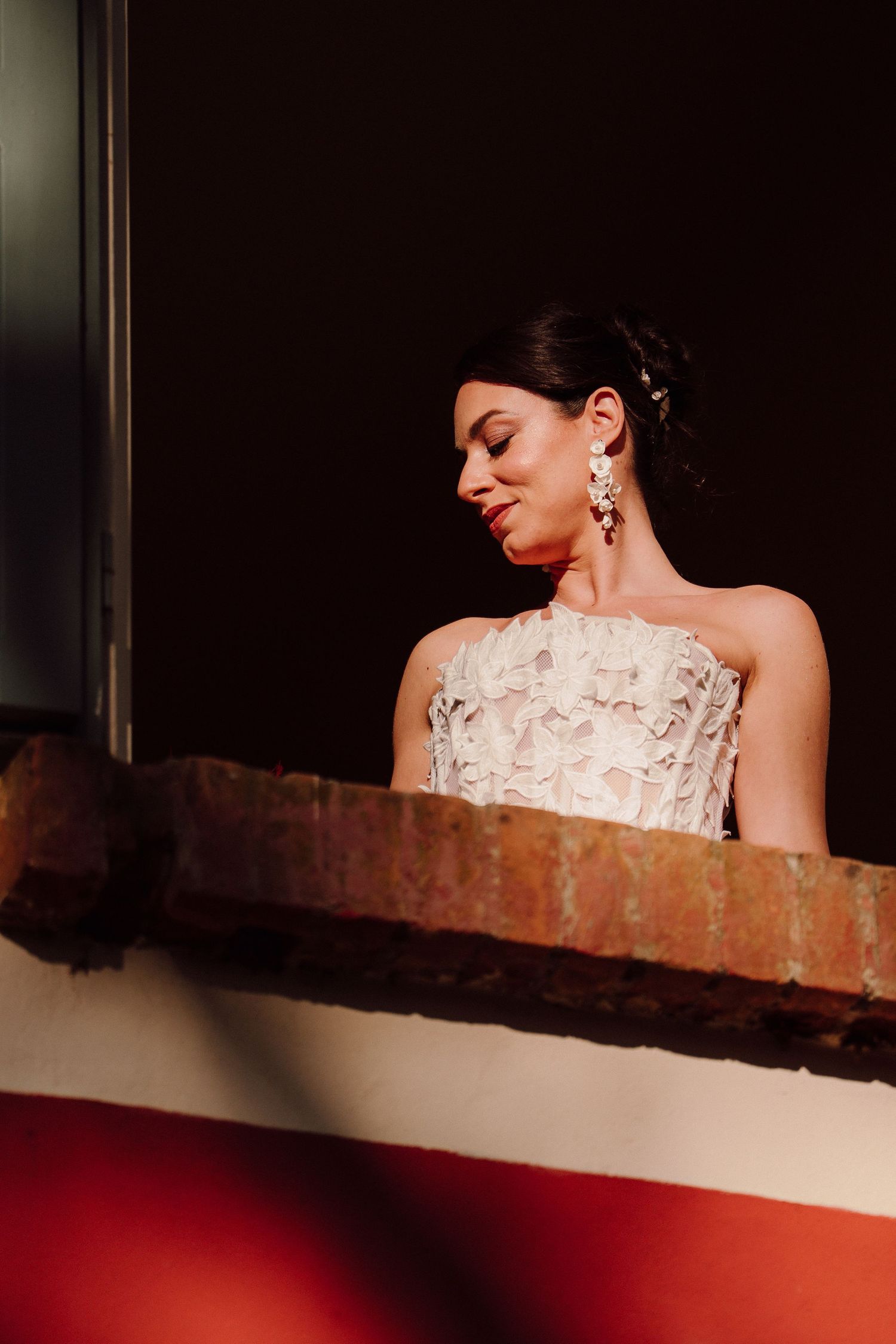 Bride in strapless embellished gown gazes down while standing against a brick wall at dusk.