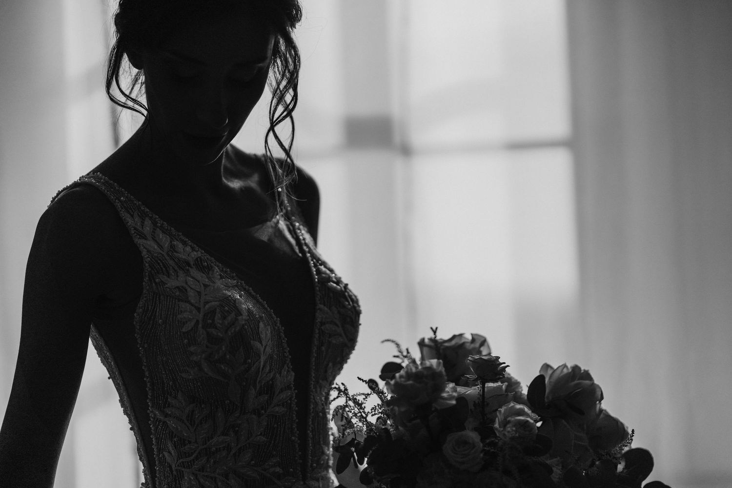 Silhouette of bride in embellished gown holding bouquet against window light.