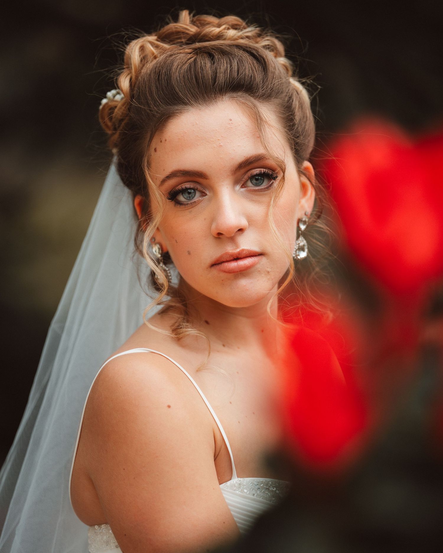 Bridal portrait with delicate veil and elegant makeup against blurred red flowers.