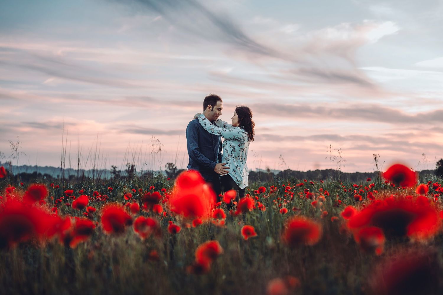 A couple embraces in a stunning poppy field at sunset with red flowers in the foreground.