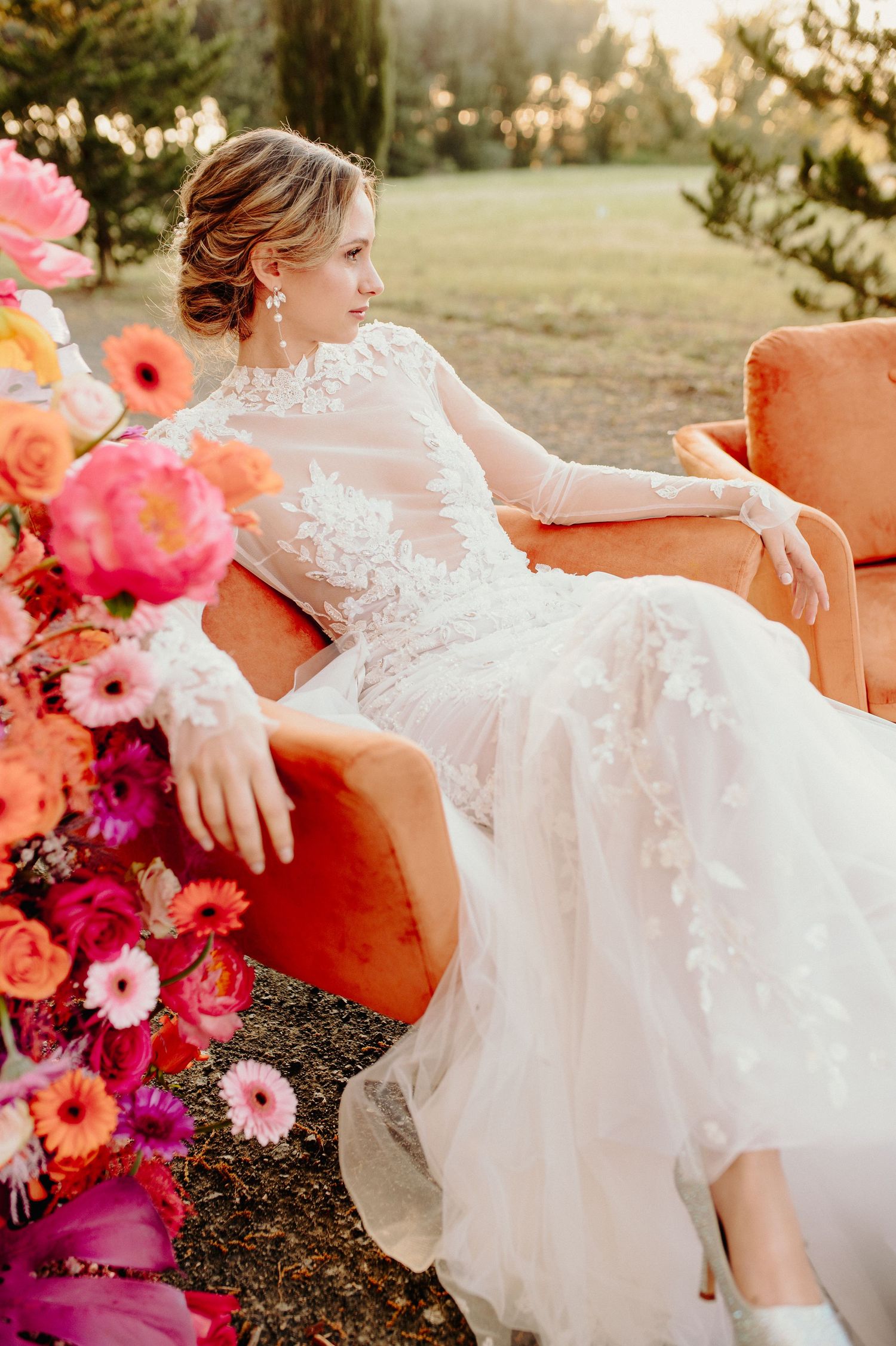 Bride in lace gown reclining on orange velvet chair amid colorful floral arrangements.