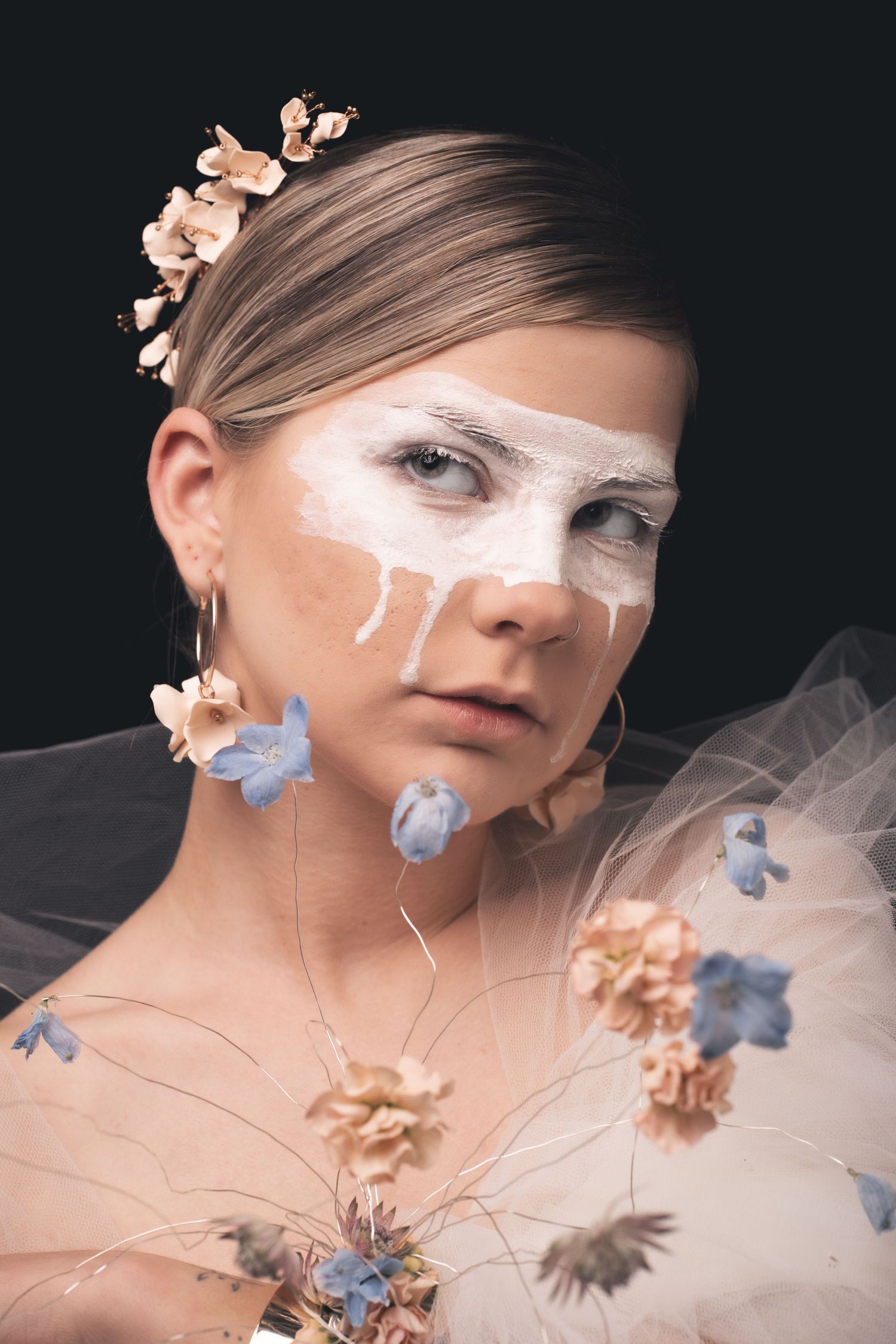 Artistic portrait with white face paint and delicate peach and blue flowers against a dark background.