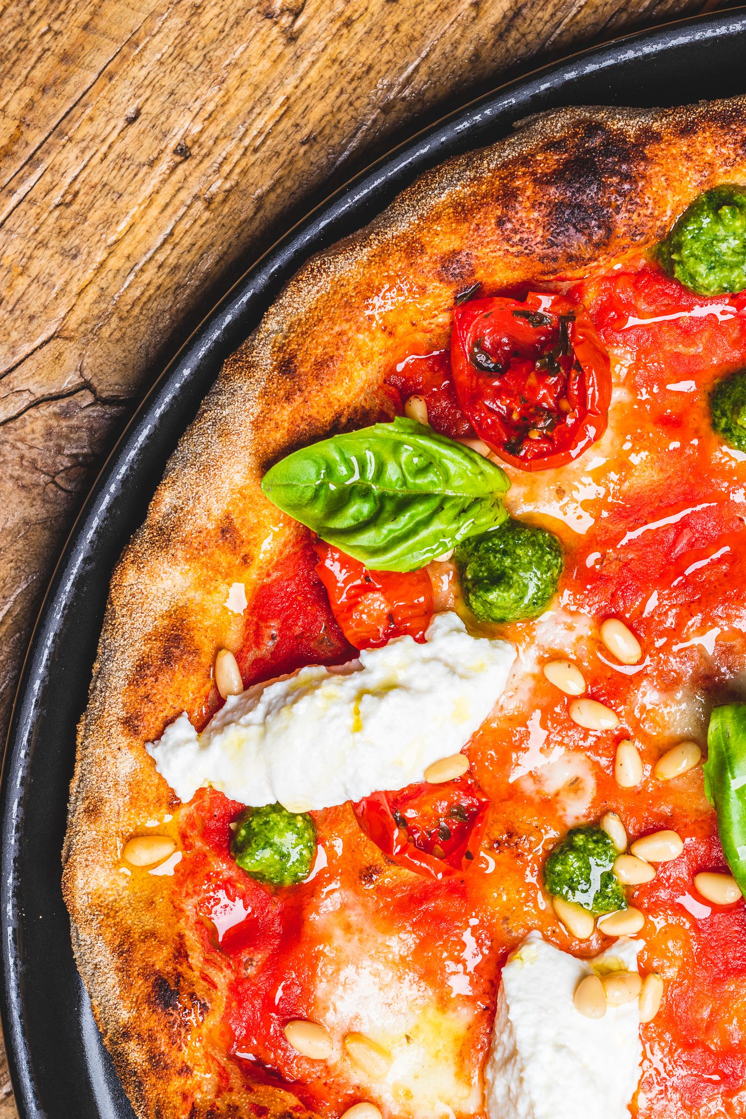 Wood-fired pizza with fresh burrata, tomatoes, pine nuts and basil leaves on dark surface.