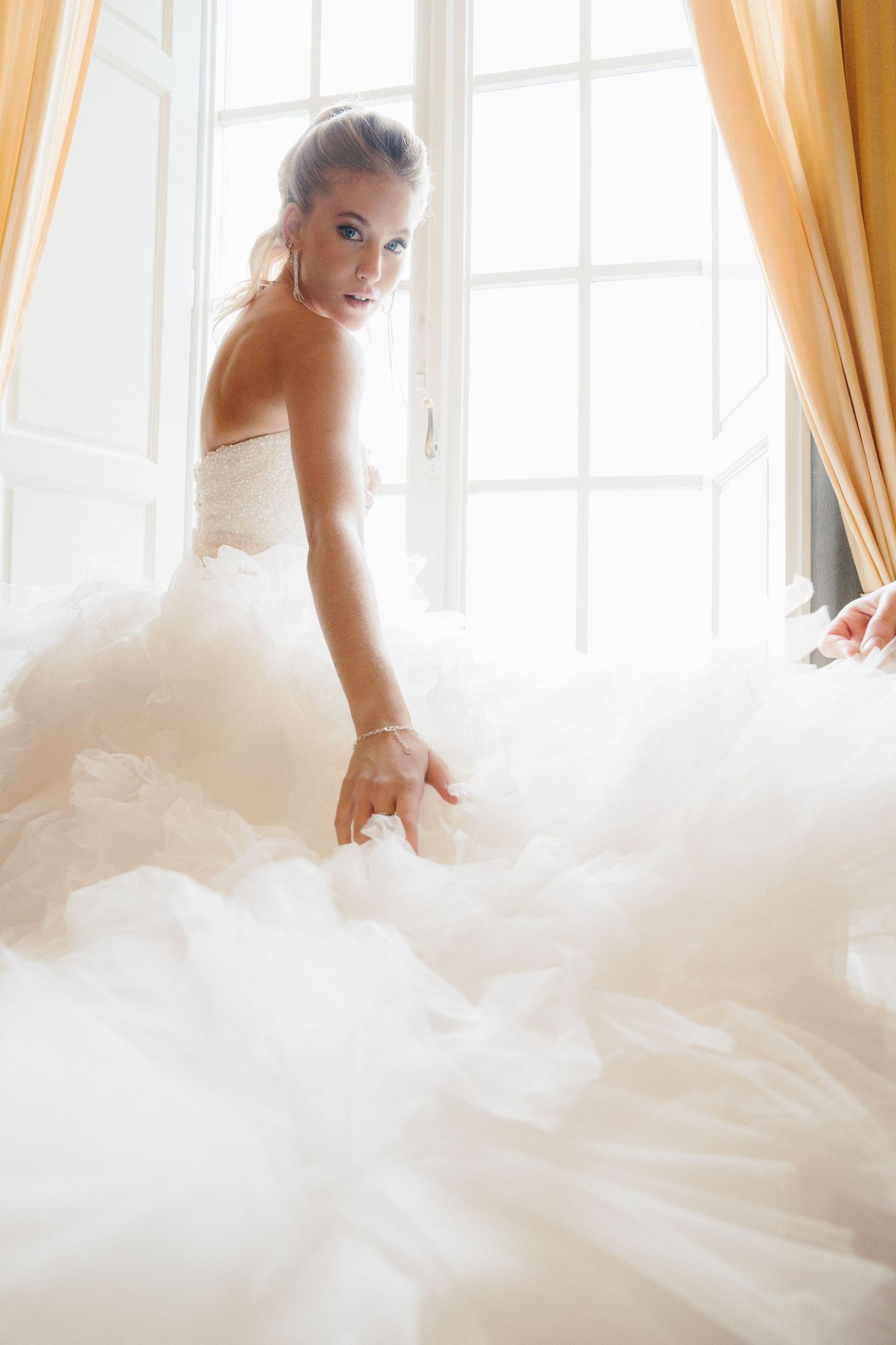Dreamy bridal portrait with flowing tulle dress near sun-lit window.