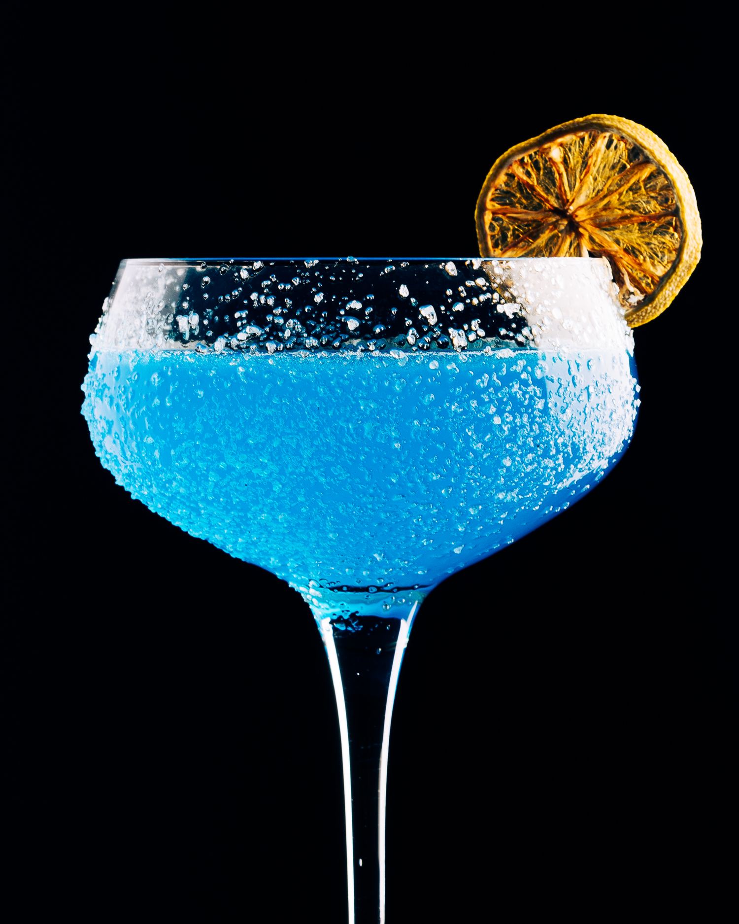 Bright blue cocktail in a coupe glass garnished with dehydrated citrus against black background.