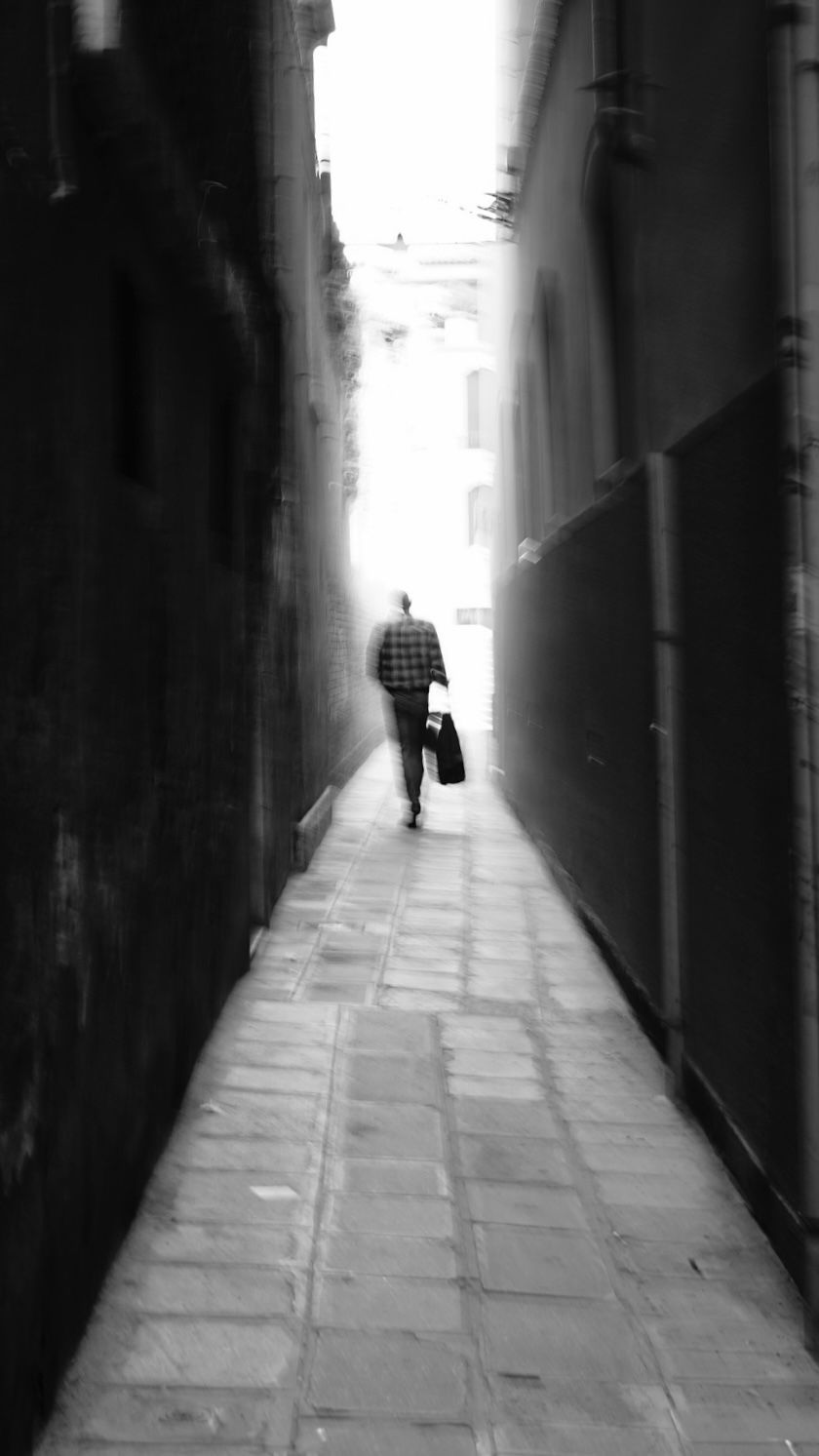 Black and white photo of person walking through narrow alleyway between tall buildings.