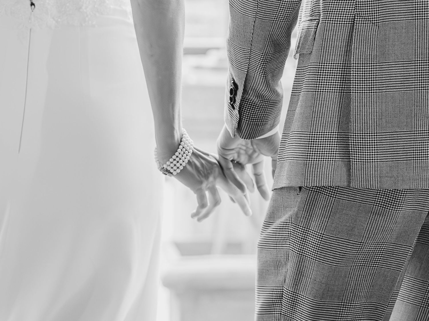 An intimate black and white detail shot of a couple holding hands, showing wedding attire.