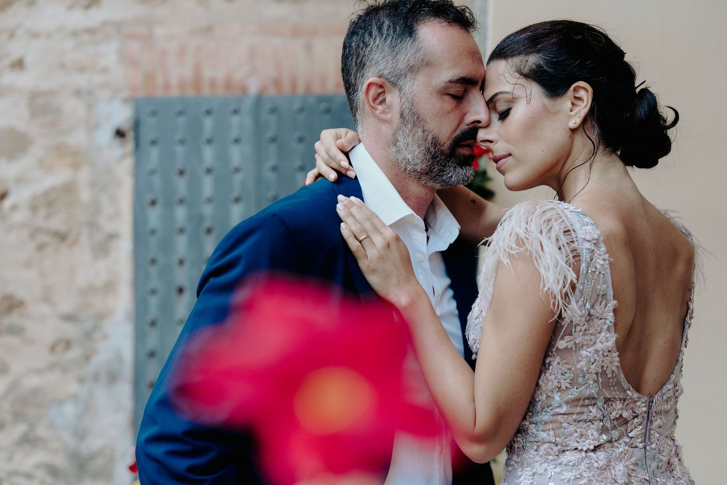 Romantic wedding couple shares an intimate moment near rustic stone wall with red blur in foreground.
