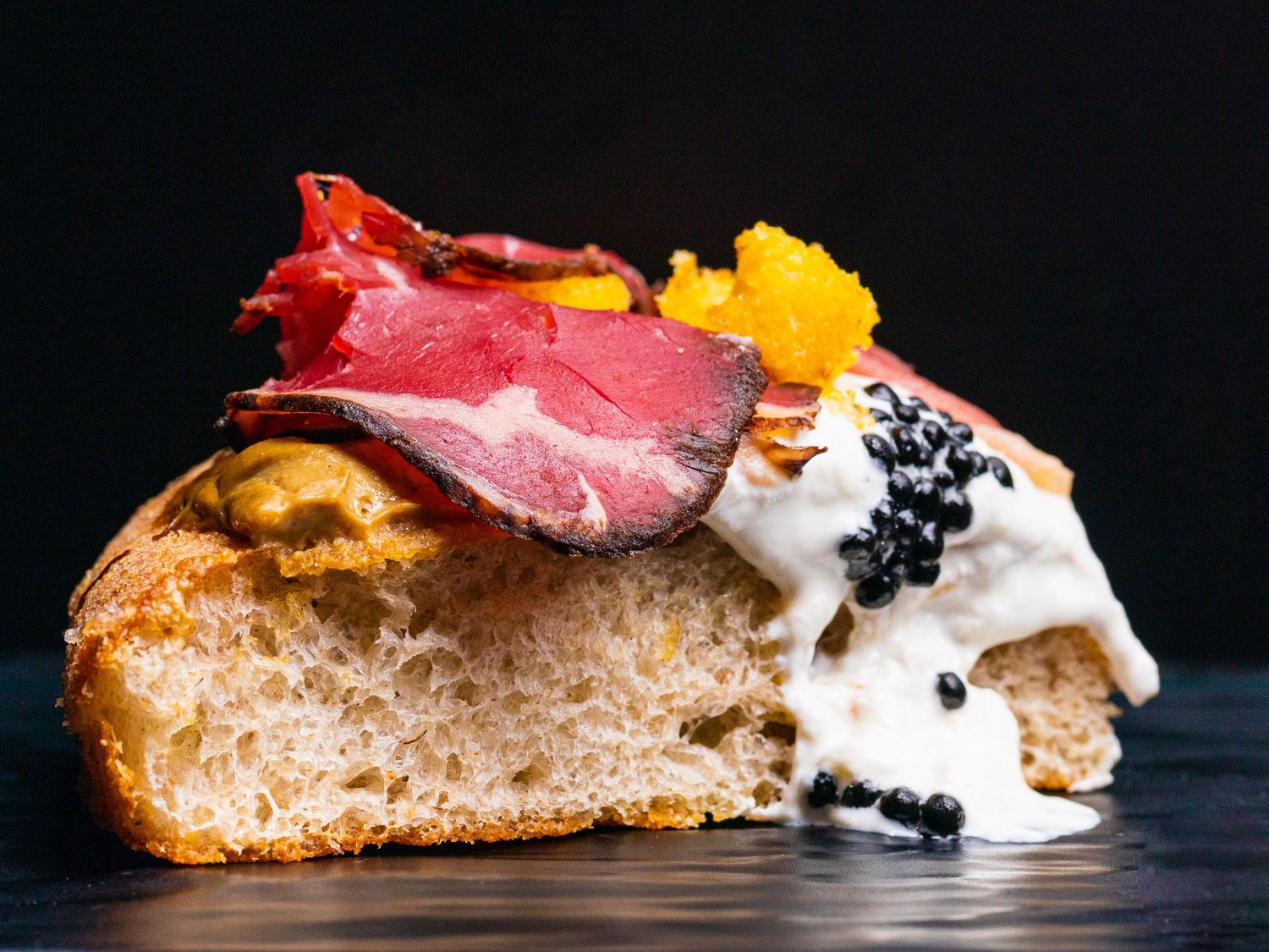 Artfully plated bruschetta topped with prosciutto, burrata cheese, caviar and golden garnish on dark surface.