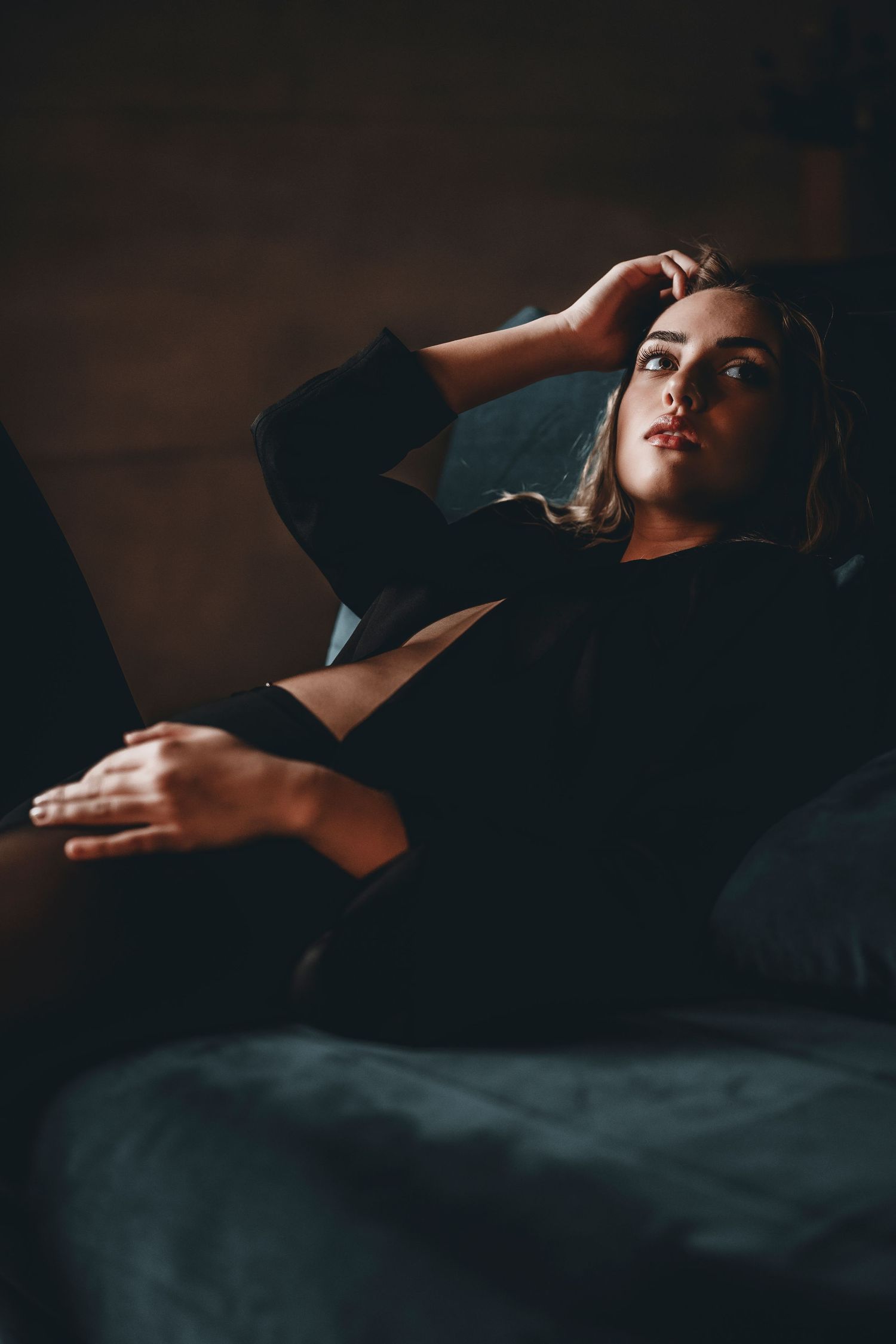 Moody portrait of a person reclining on dark furniture in dramatic lighting.