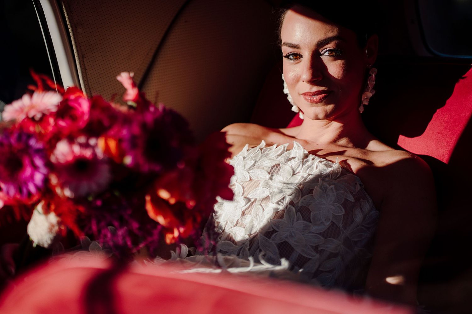 Bride in detailed white gown sits in car with vibrant red and pink floral bouquet.