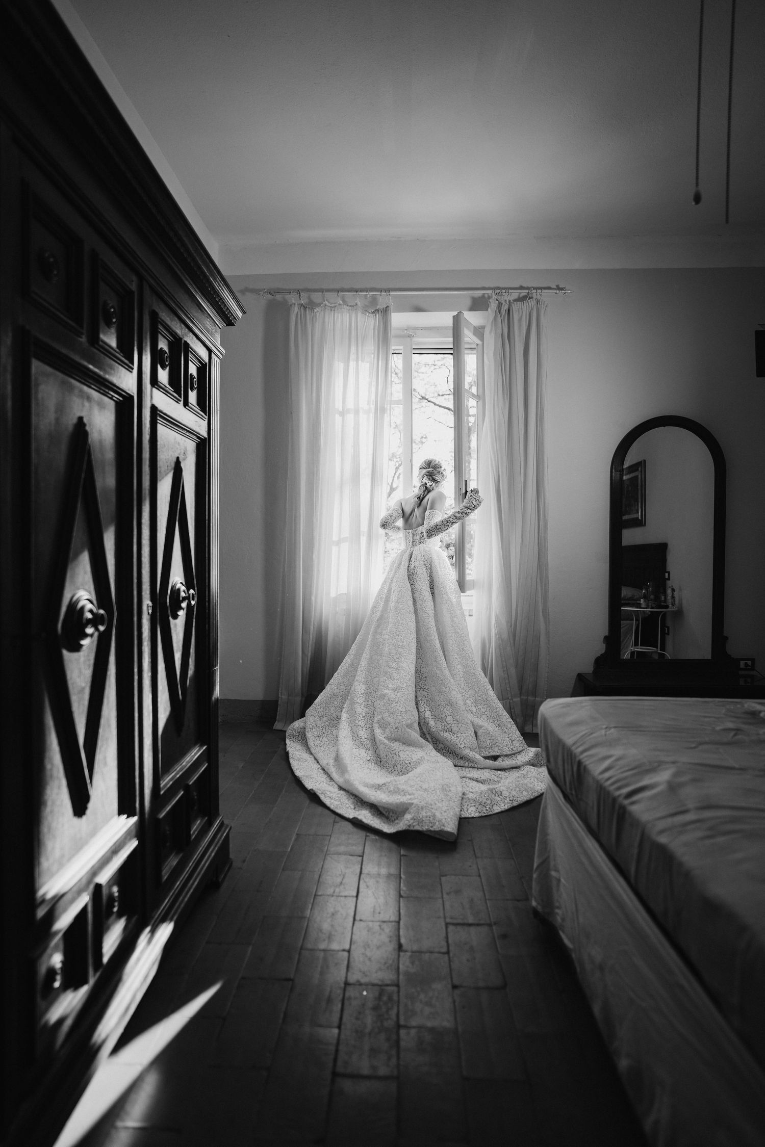 Elegant wedding dress hanging by sunlit window in vintage bedroom with hardwood floors.