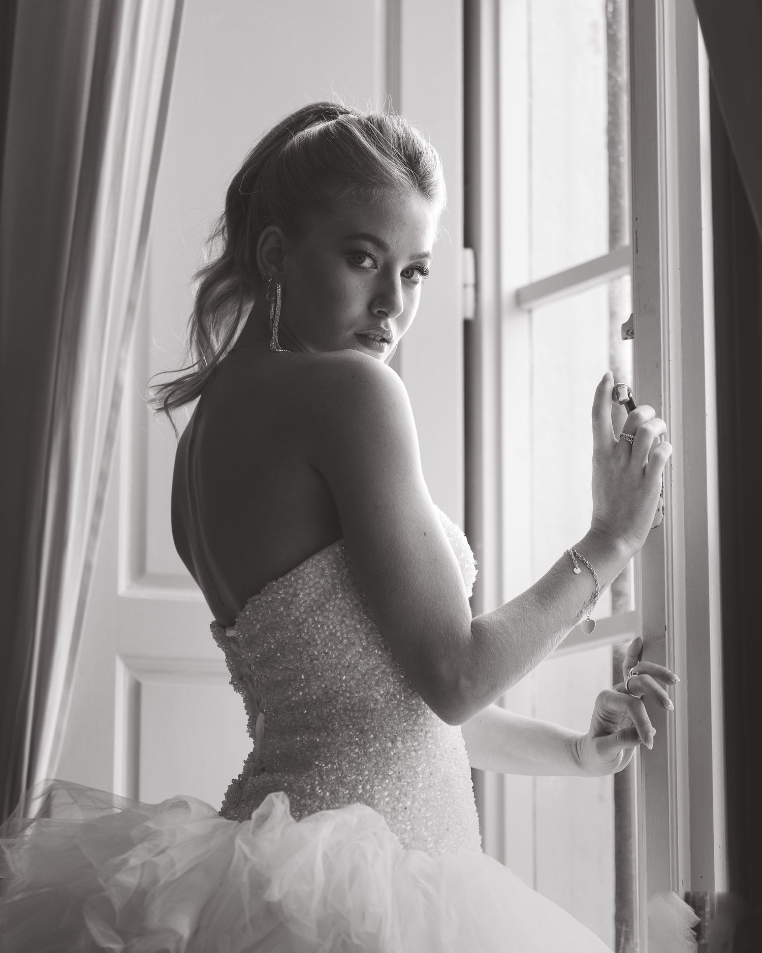 Bride in a beaded ballgown looks over her shoulder while standing by a bright window.