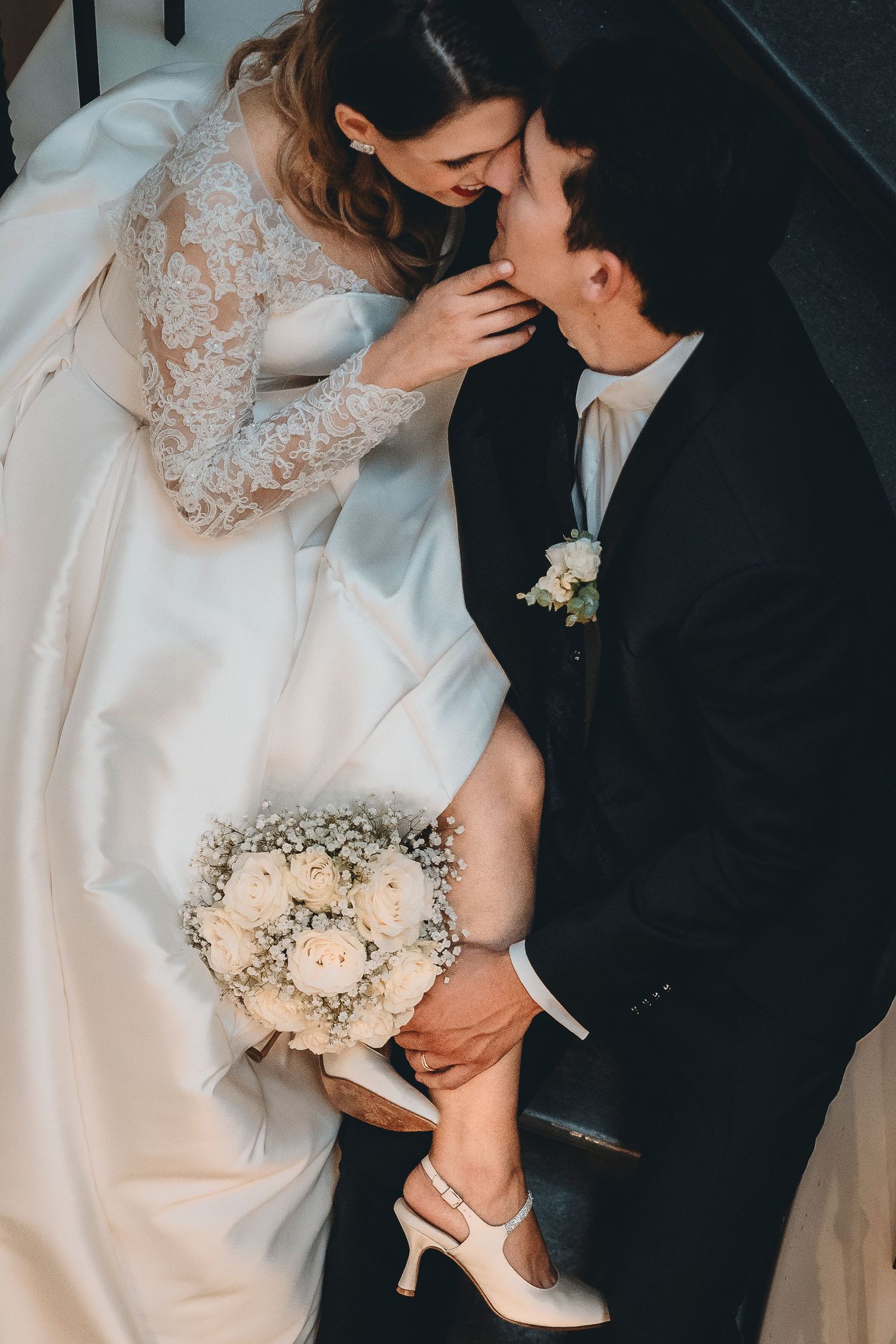 Romantic wedding couple sharing an intimate moment with bride holding bouquet and showing vintage heels.