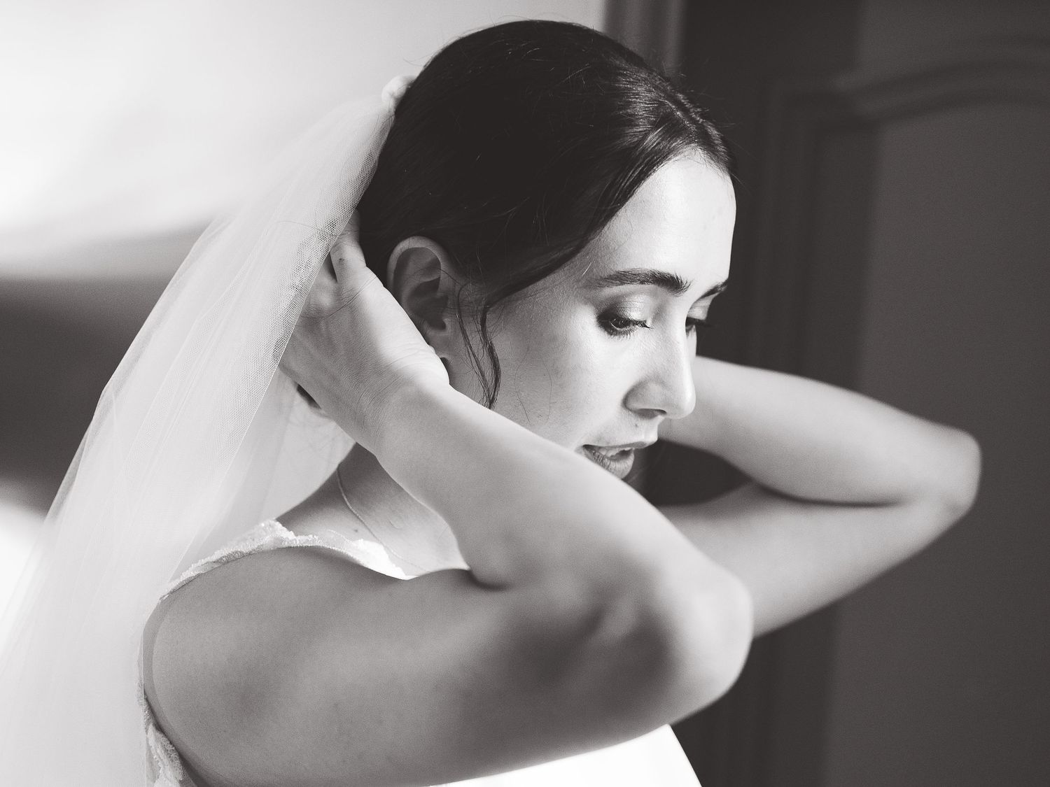 A black and white photo of a bride adjusting her veil with soft natural light.
