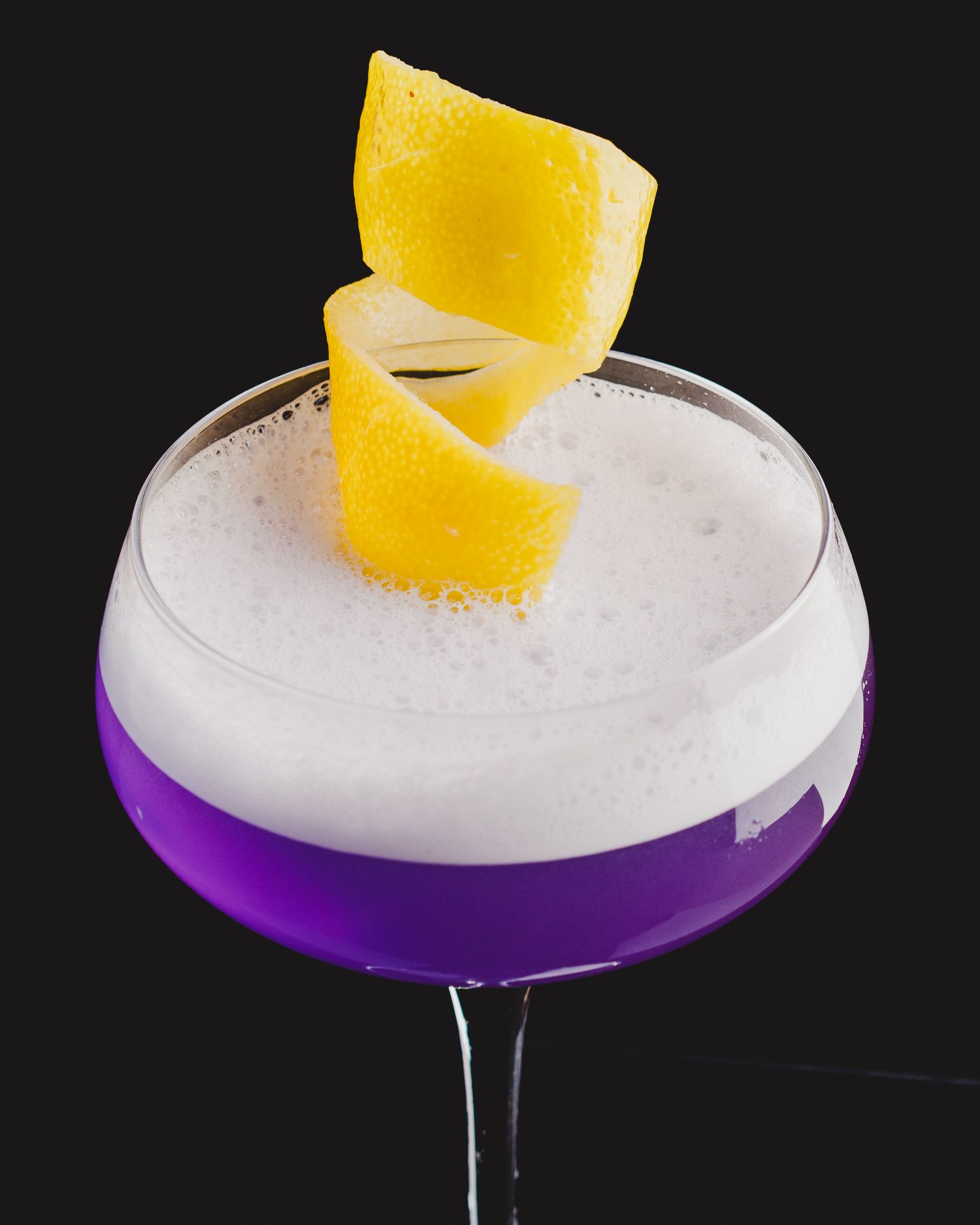 Purple cocktail with white foam and twisted lemon garnish in a coupe glass.