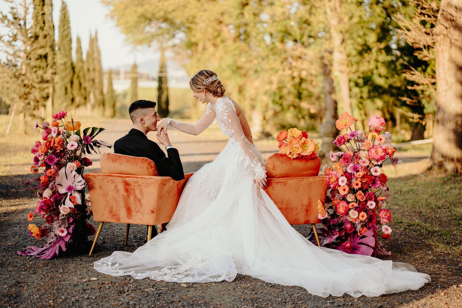 Romantic wedding scene with couple and orange floral arrangements on rustic bench in outdoor garden setting.