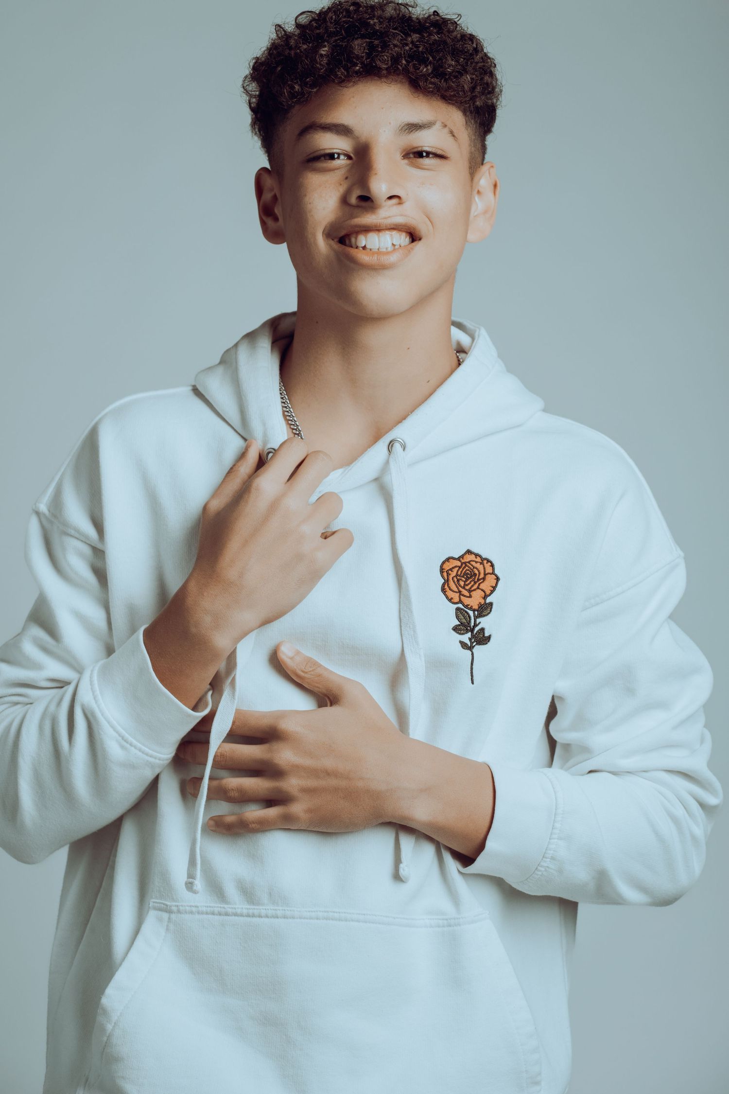 Portrait of person in white hoodie with flower emblem smiling against gray background.