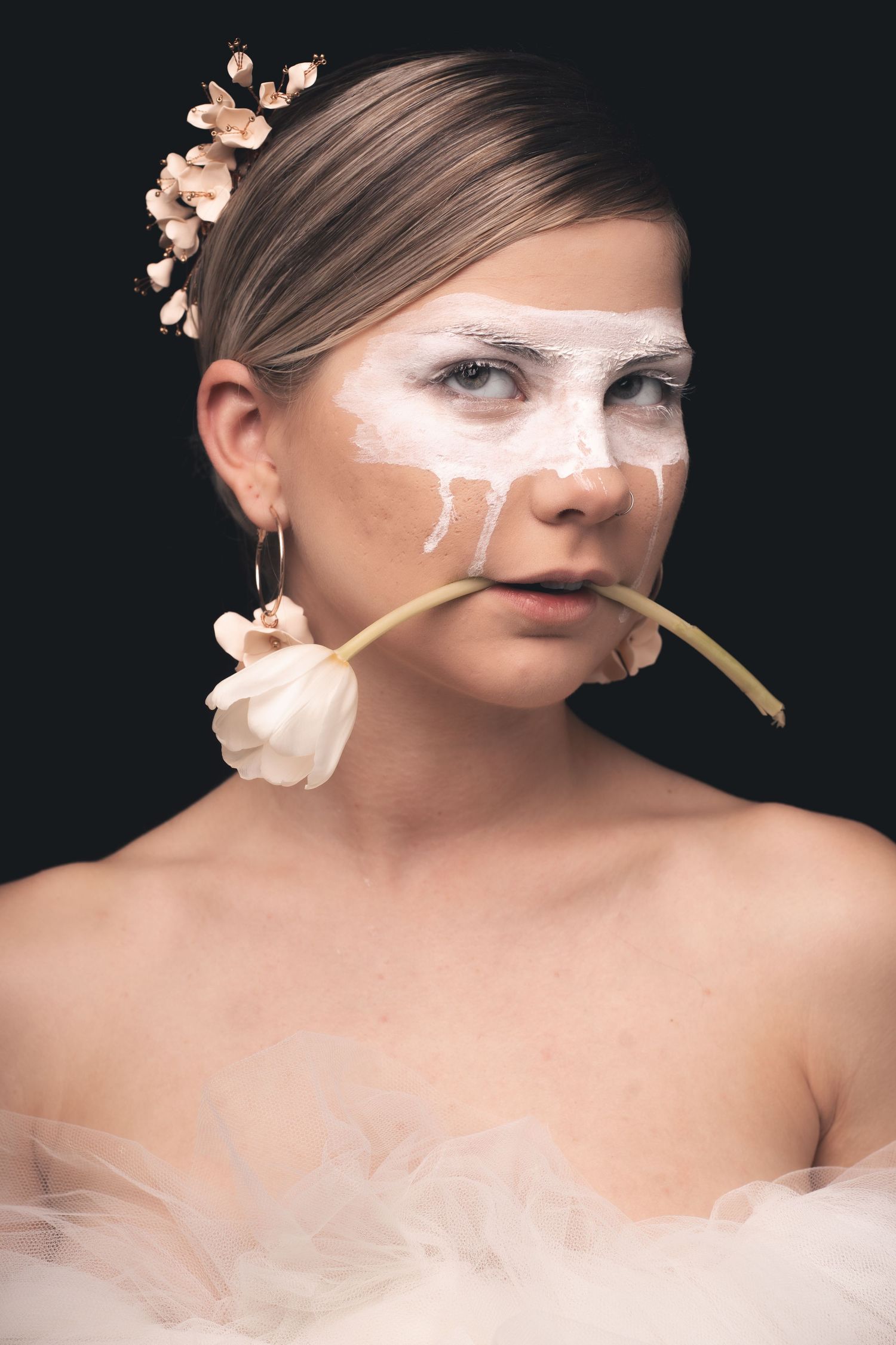 Creative portrait featuring white face paint, dried flowers, and a single stem held between lips.