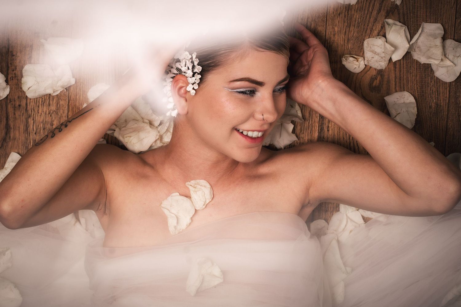 A radiant smiling figure lying among delicate white rose petals against a rustic wooden background.