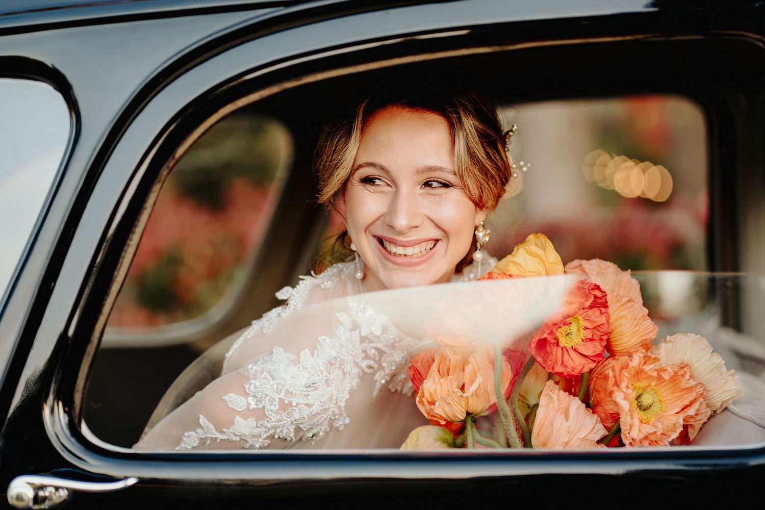 A bride wearing a lace dress smiles while seated in a vintage car with beautiful orange and pink roses.