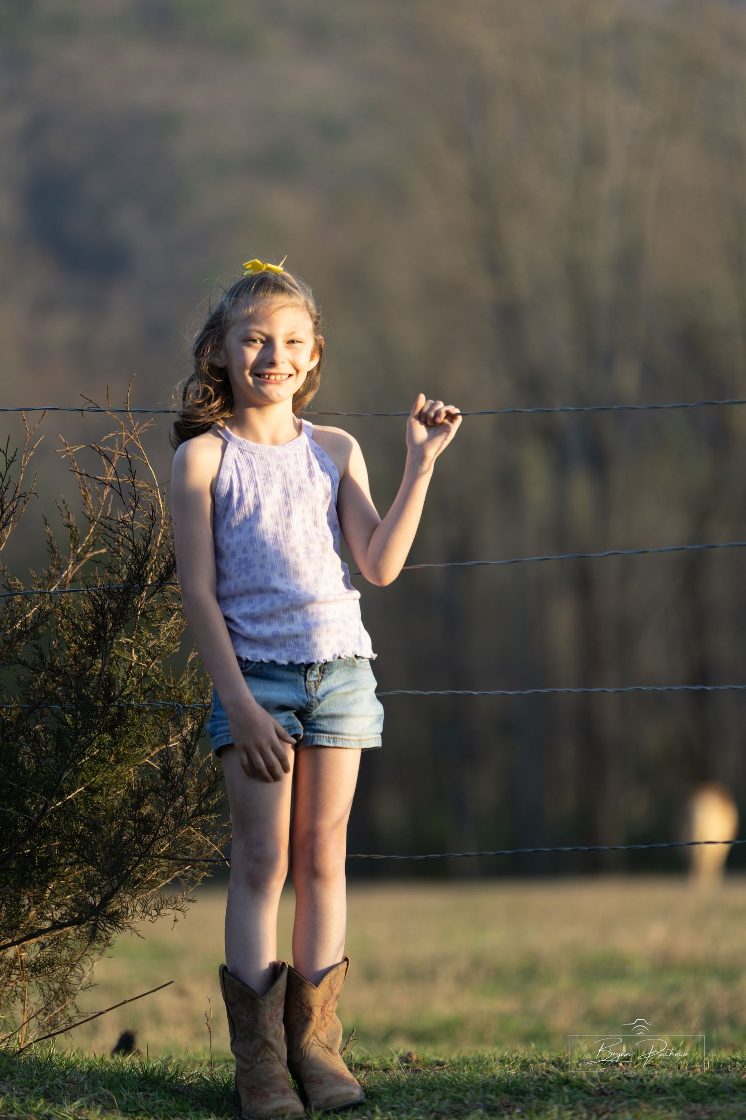 Child in a white top and denim shorts poses playfully by a wire fence wearing brown cowboy boots on a sunny day.