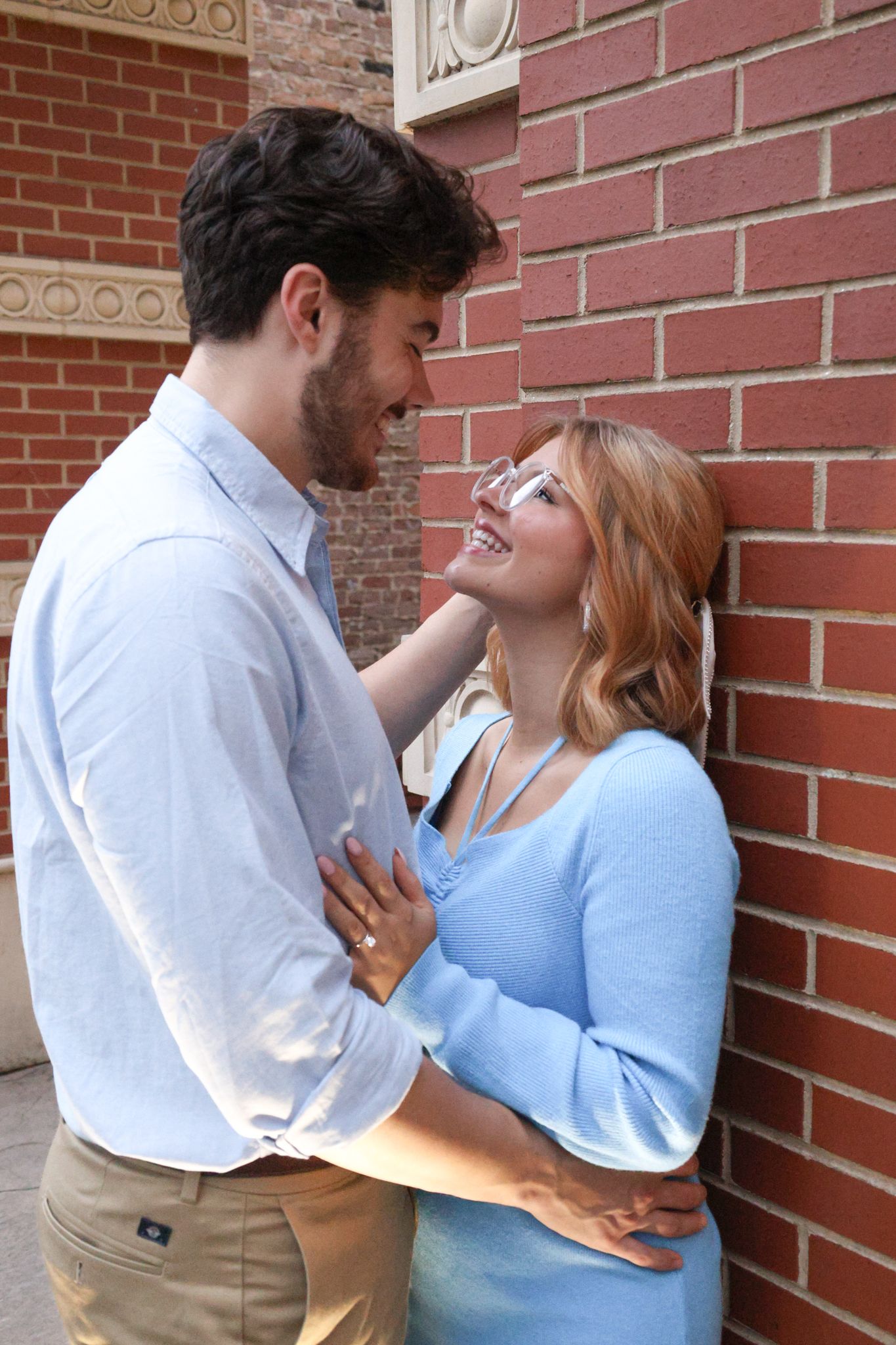 A romantic couple embraces against a brick wall, sharing an intimate moment in casual attire.