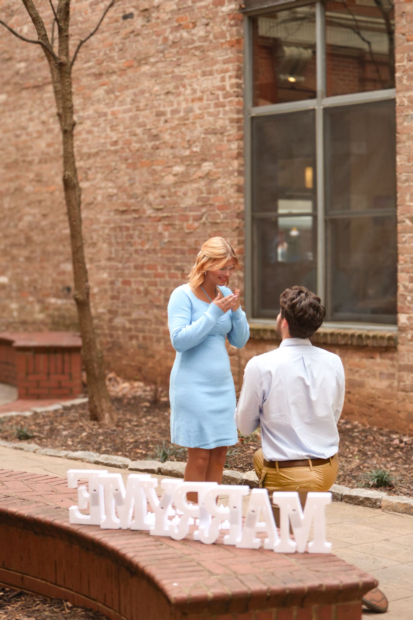 Proposal moment captured with 'Marry Me' sign on brick ledge during surprise engagement.