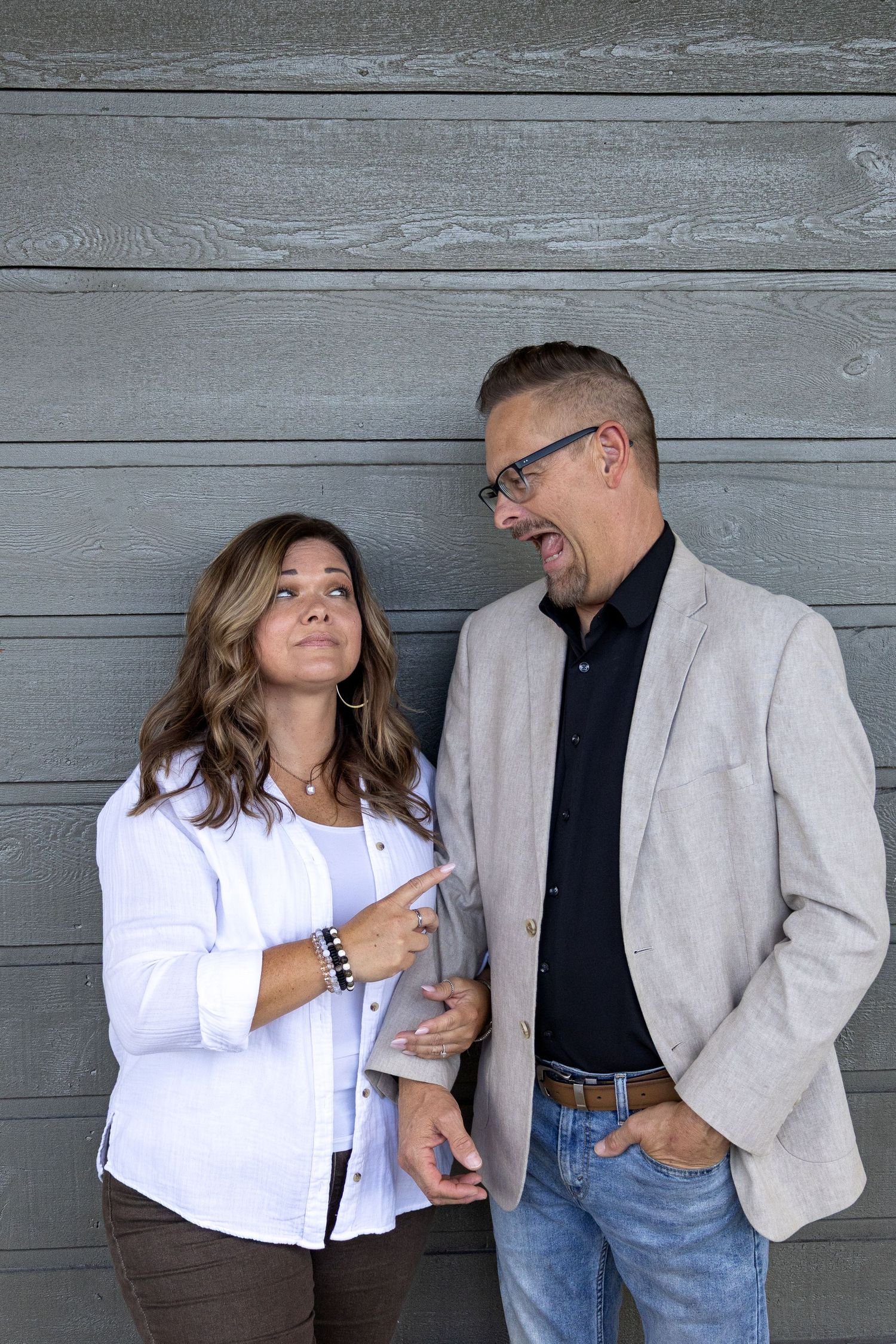 Two people in casual business attire share a lighthearted moment against a gray wooden wall background.
