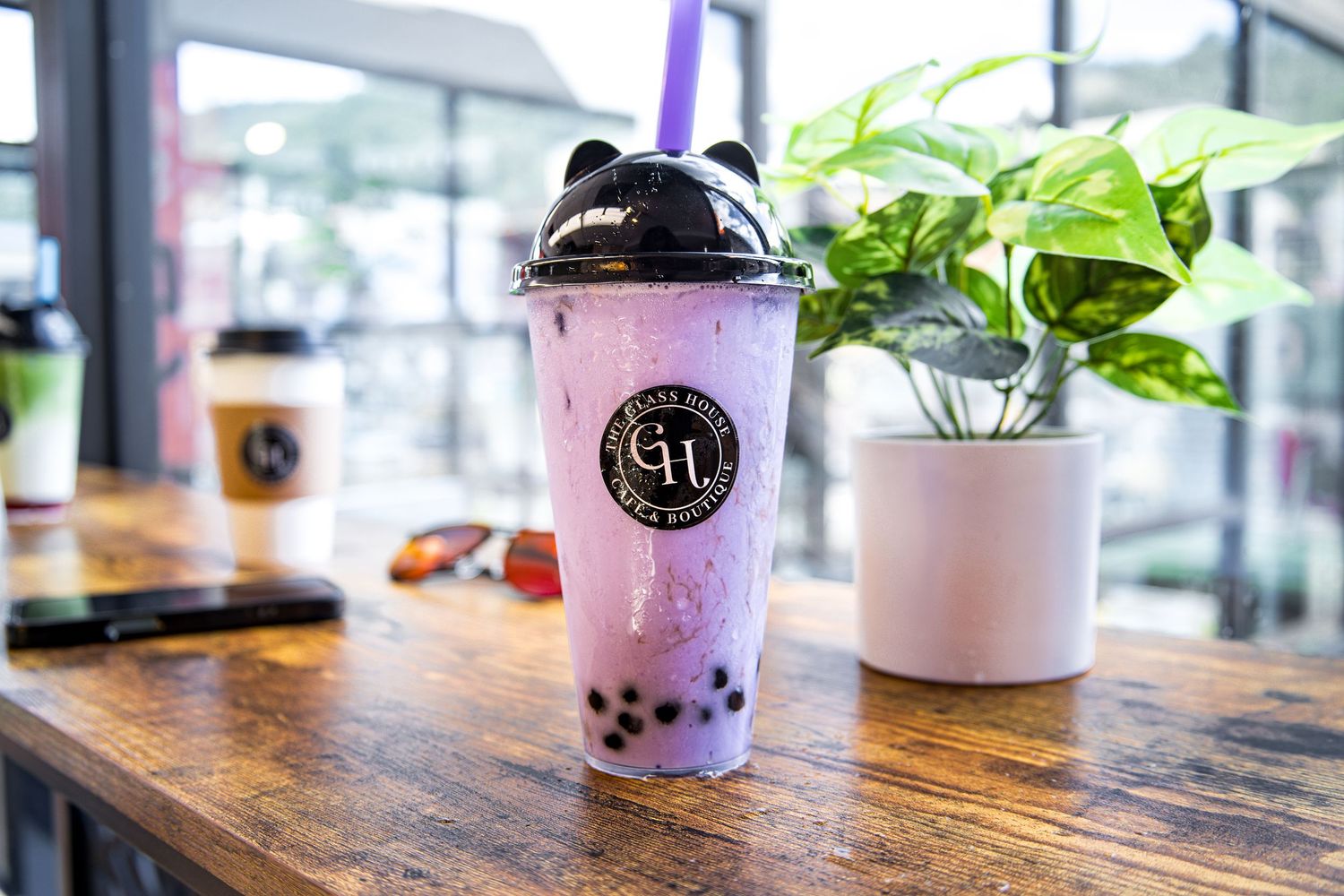 Purple bubble tea drink with black boba pearls in branded cup sitting on wooden table with plant decor.